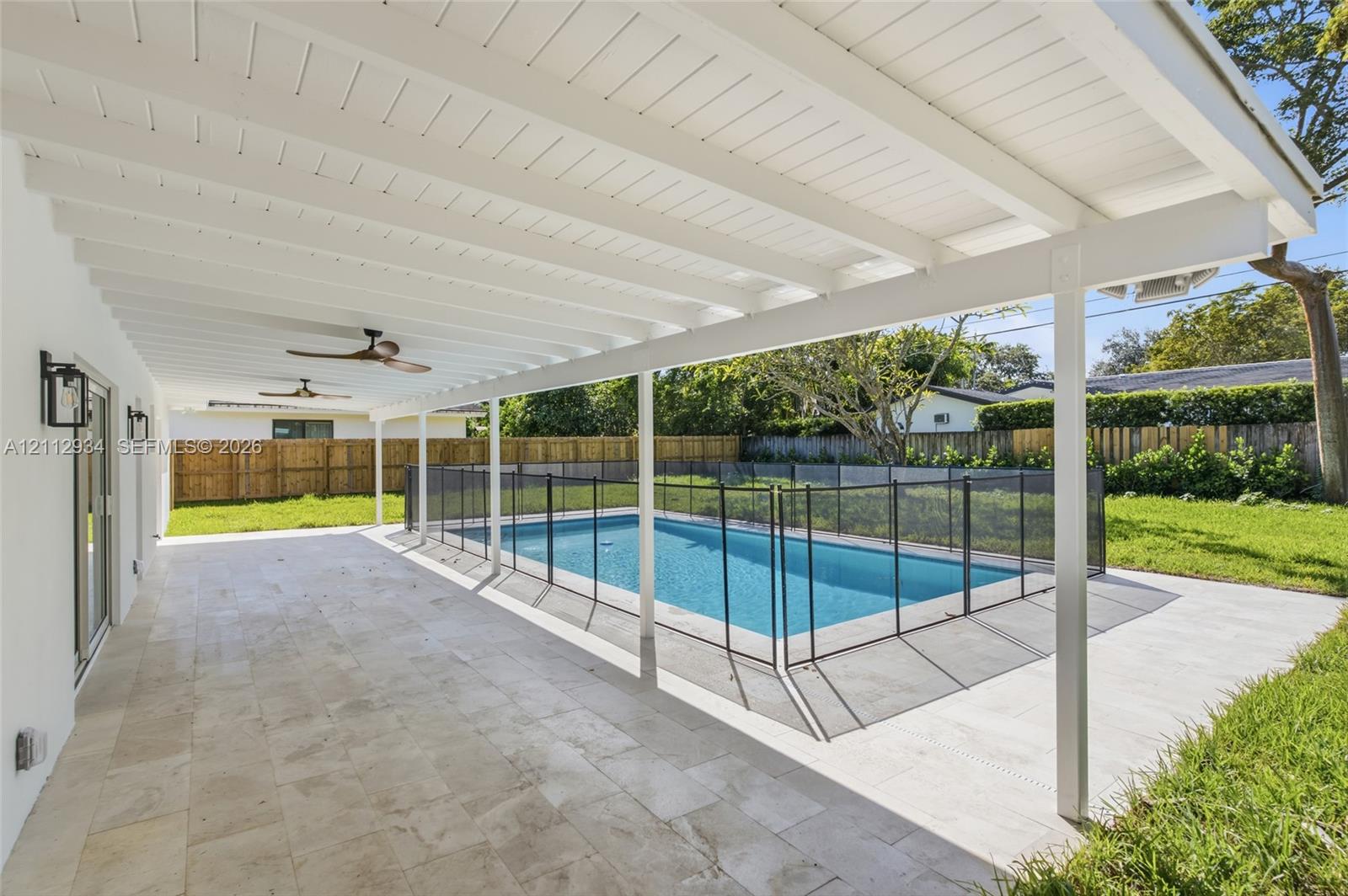 Covered patio featuring stone-tiled flooring and ceiling-mounted fans, overlooking a swimming pool with an installed mesh safety fence and a fenced backyard