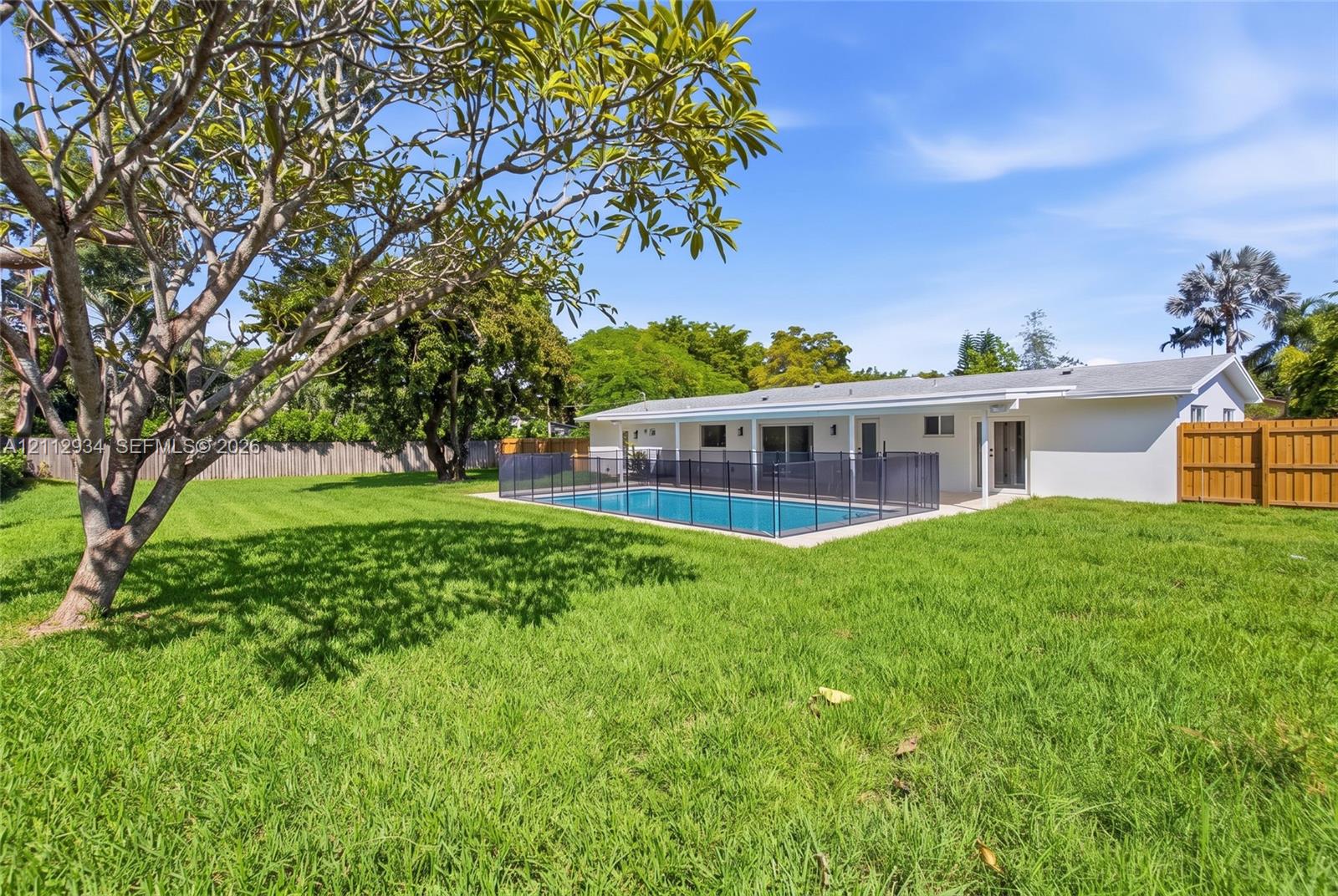 Spacious backyard featuring a grass lawn, mature trees, a swimming pool with a perimeter safety fence, a covered patio, and wood-style privacy fencing
