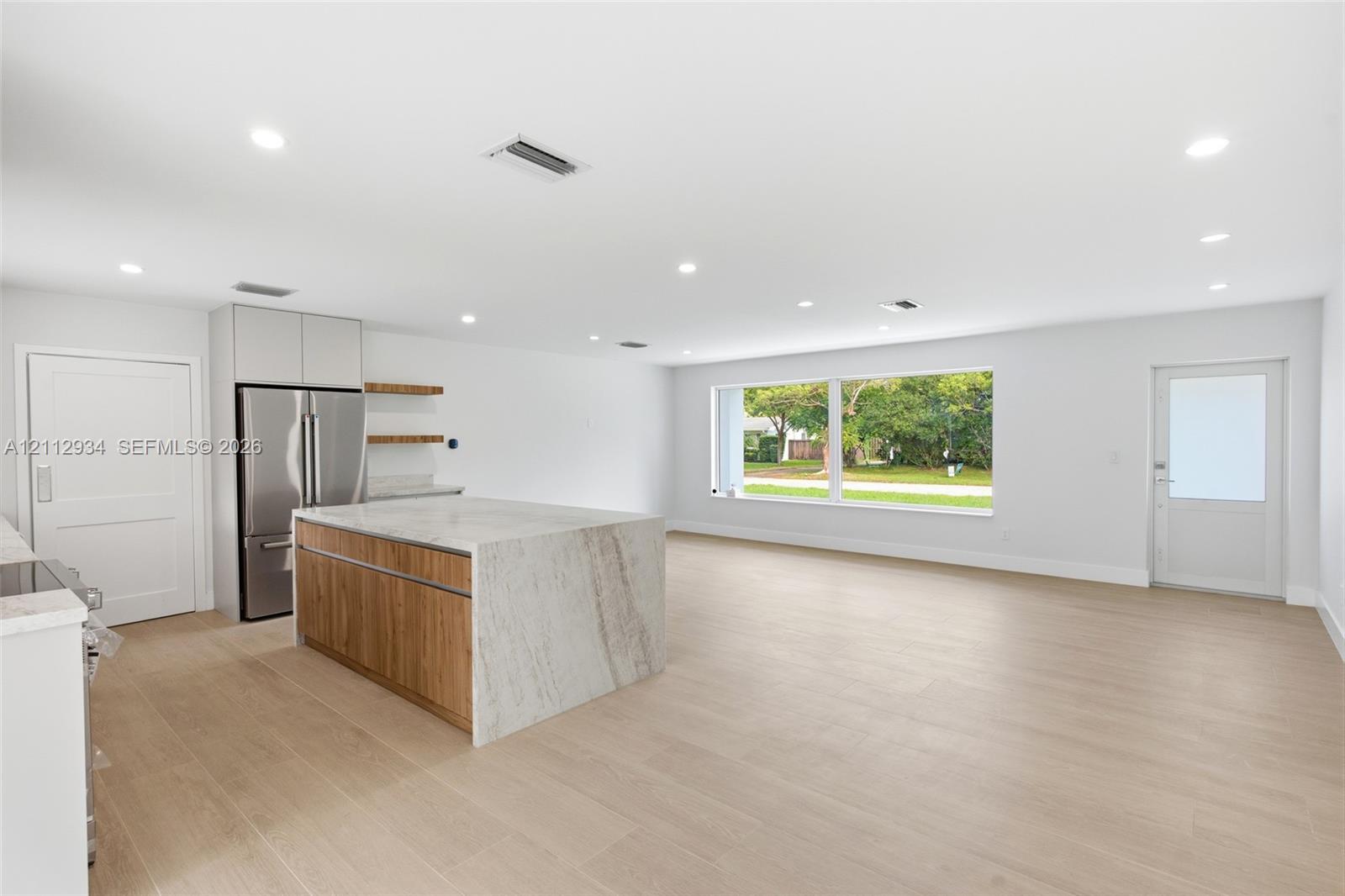 Open-concept kitchen featuring an island with stone-finish waterfall countertops and wood-finish cabinetry, stainless steel refrigerator, recessed lighting, and wood-finish flooring throughout