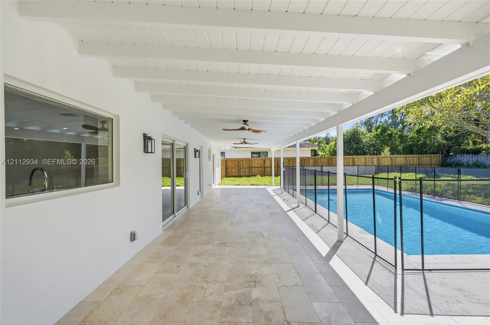 Covered patio featuring natural stone tile flooring, exposed beam ceiling with dual mounted fans, sliding glass doors, and a pool with perimeter fencing