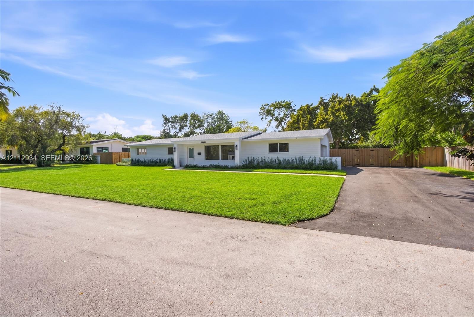 Single-story ranch exterior featuring a white stucco facade, expansive front lawn, asphalt driveway, and wood-panel perimeter fencing