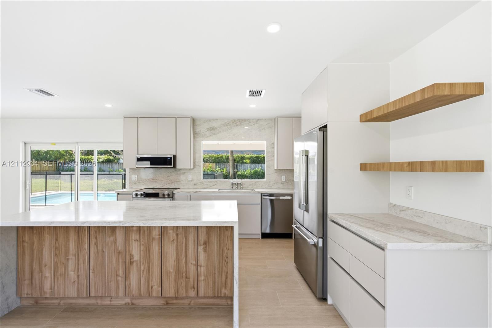 Kitchen featuring a wood-tone island with waterfall stone countertop, neutral flat-panel cabinetry, stone backsplash, built-in stainless steel appliances, and floating wood shelving