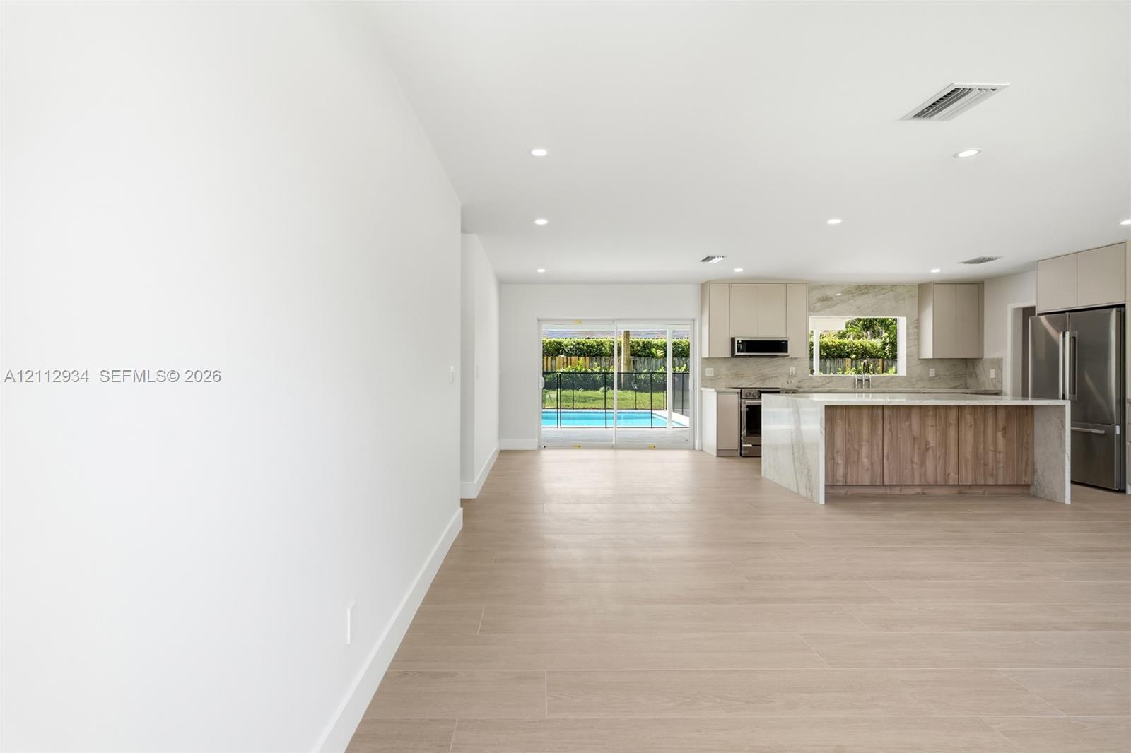 Open-concept kitchen featuring an island with a waterfall-edge stone countertop, wood-finish cabinetry, recessed lighting, and wood-finish flooring, with sliding glass doors leading to a pool area