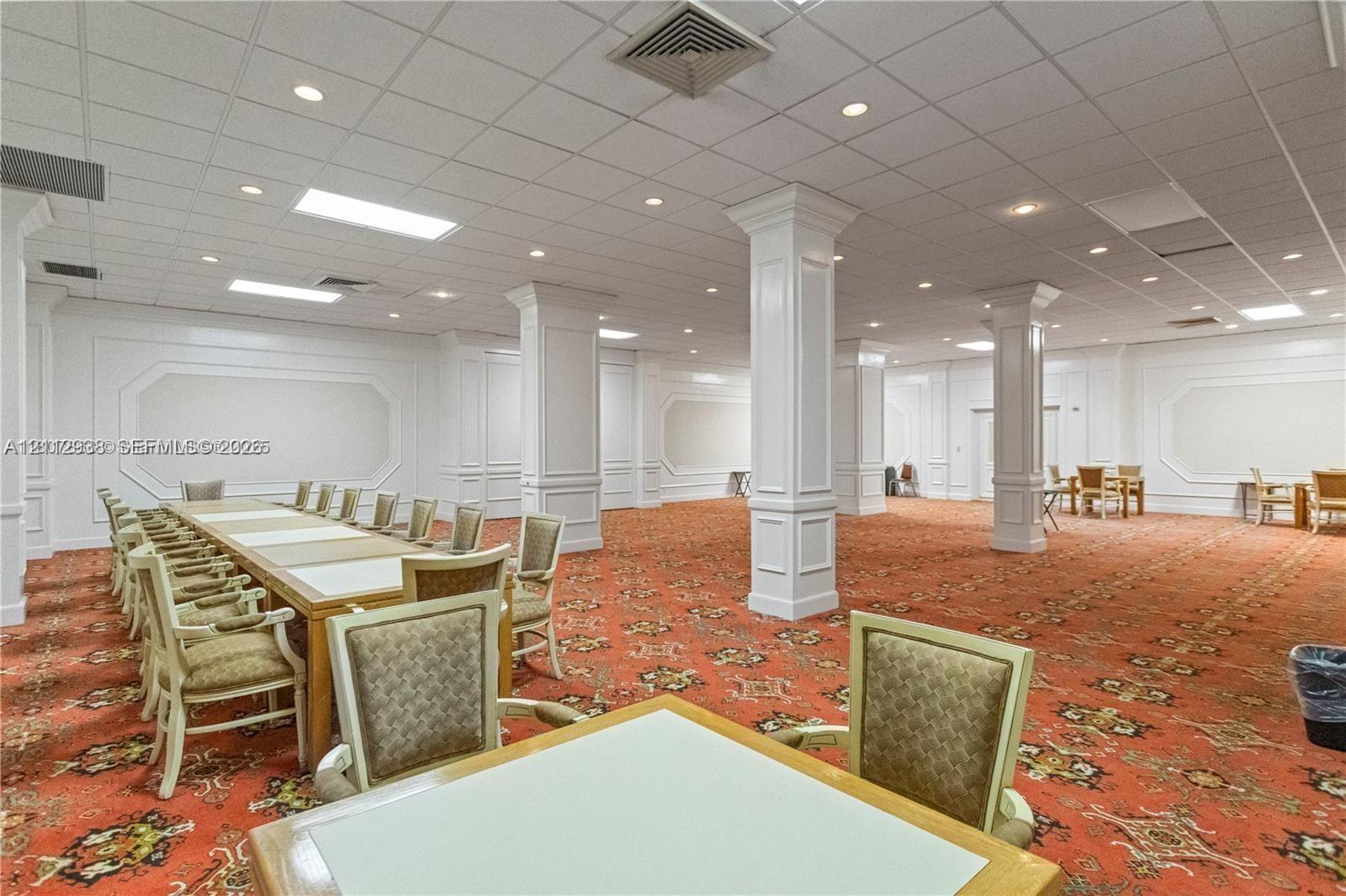 Expansive interior space featuring a drop ceiling with recessed lighting, prominent white structural columns, wall molding details, and patterned wall-to-wall carpeting