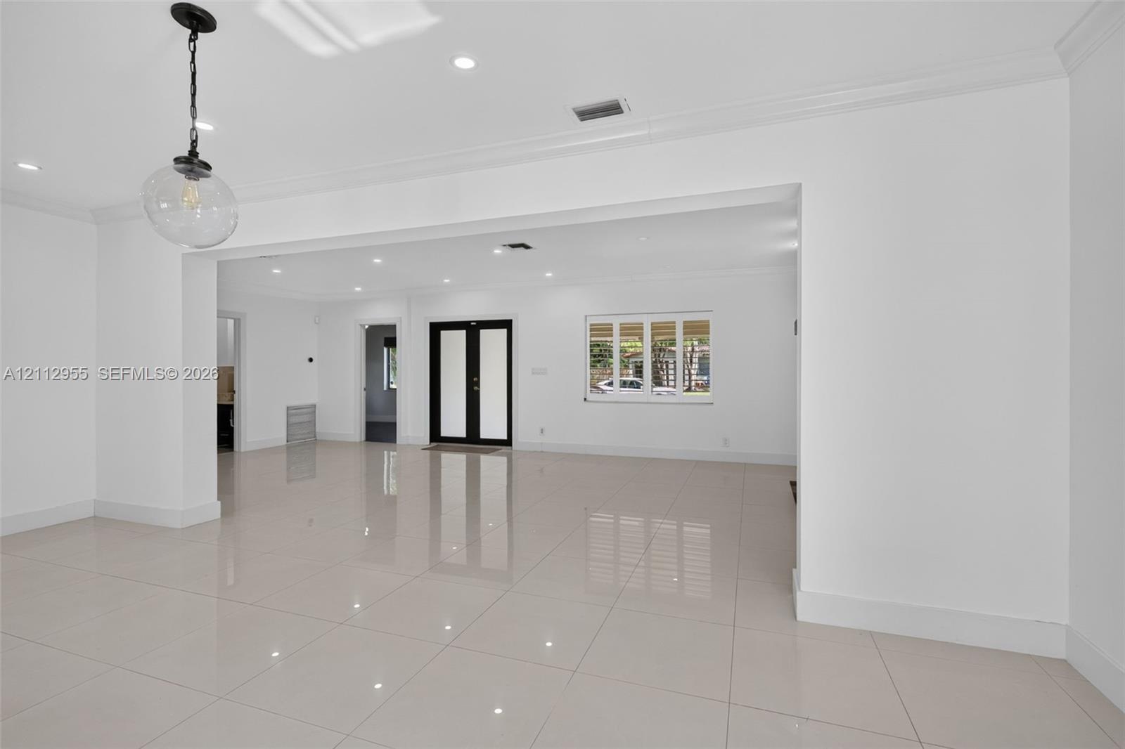 Open-concept interior featuring large-format porcelain tile flooring, crown molding, recessed lighting, double entry doors, and a decorative glass pendant light fixture
