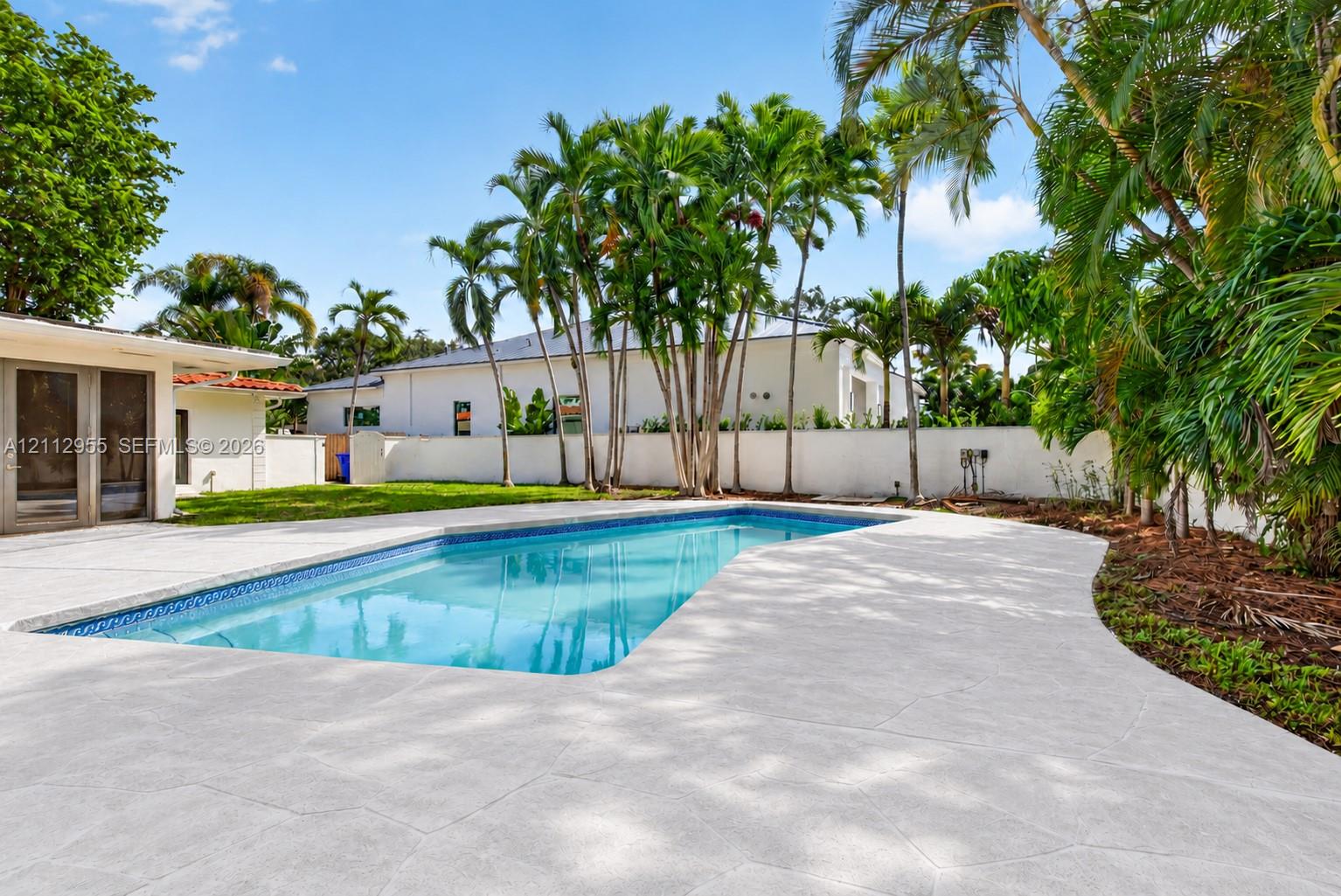 In-ground swimming pool featuring a textured concrete deck, privacy wall, manicured lawn, and mature palm trees