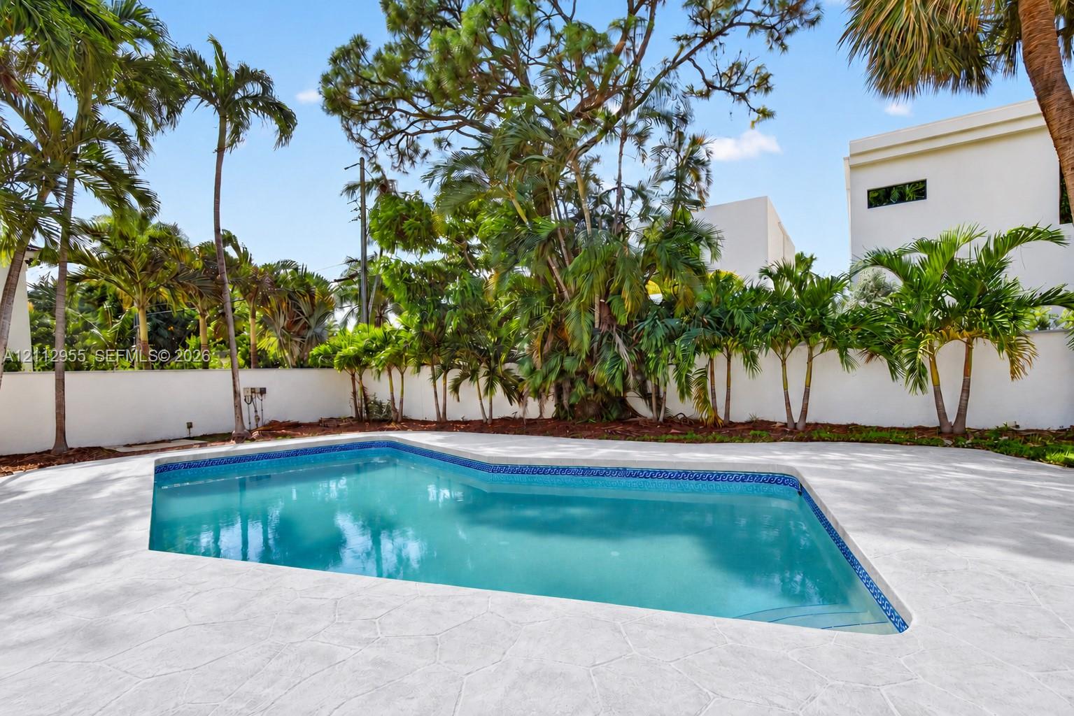 In-ground swimming pool featuring a patterned tile border, concrete deck, solid perimeter wall, and mature palm trees