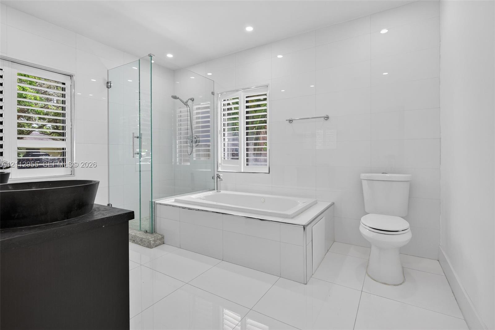 Modern bathroom featuring a glass-enclosed walk-in shower, a deep-soaking bathtub with tile surround, plantation shutters, and a dark-finish vanity with a vessel sink