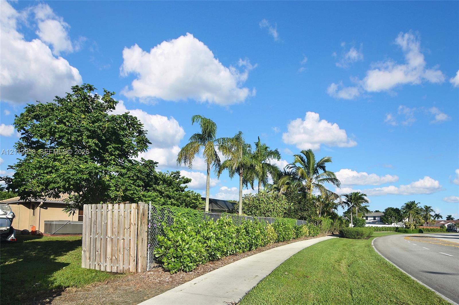 Exterior lot featuring a wooden privacy fence, chain-link perimeter, established landscaping with mature palm trees, and a concrete sidewalk