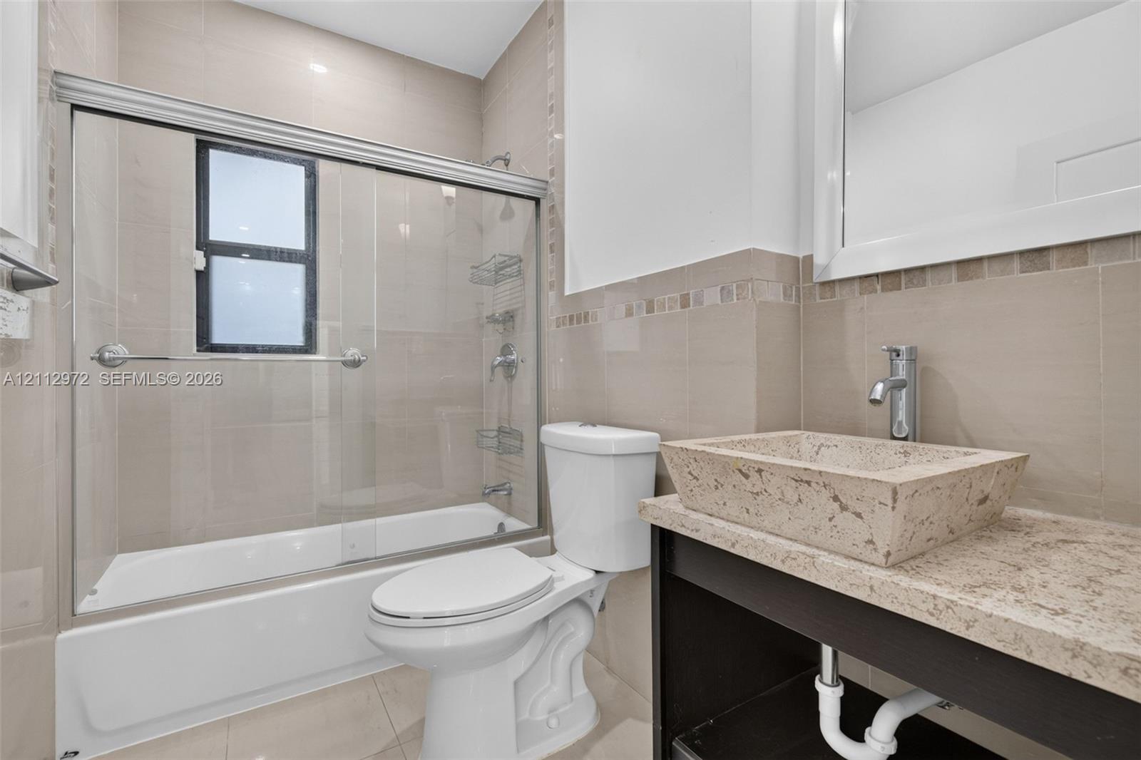 Bathroom featuring a custom stone vessel sink with a chrome faucet, solid-surface vanity countertop, glass-enclosed shower with tiled walls, and decorative accent tiling