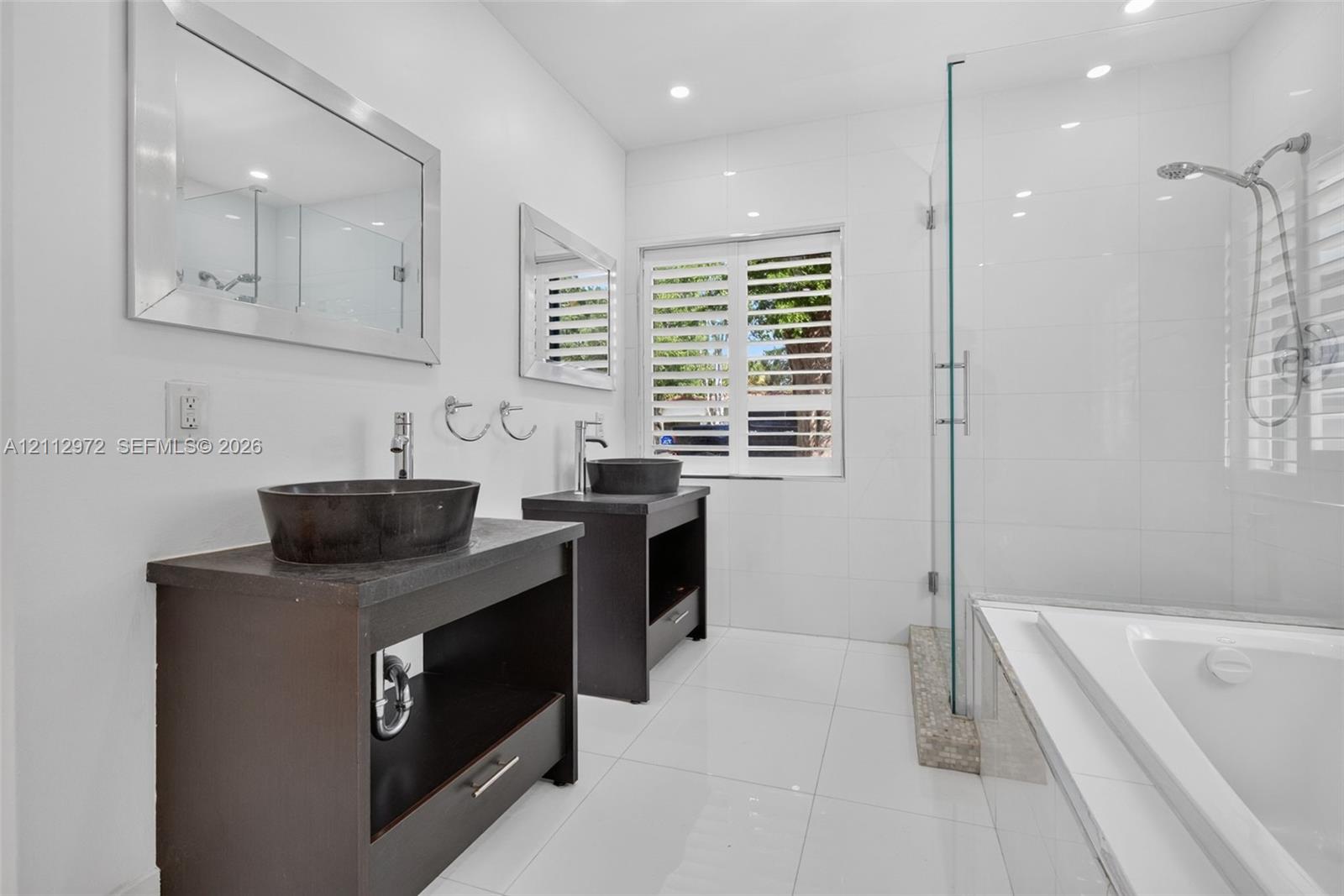 Bathroom featuring dual dark wood vanities with vessel sinks, a glass-enclosed shower, a deep soaking tub, and plantation shutters