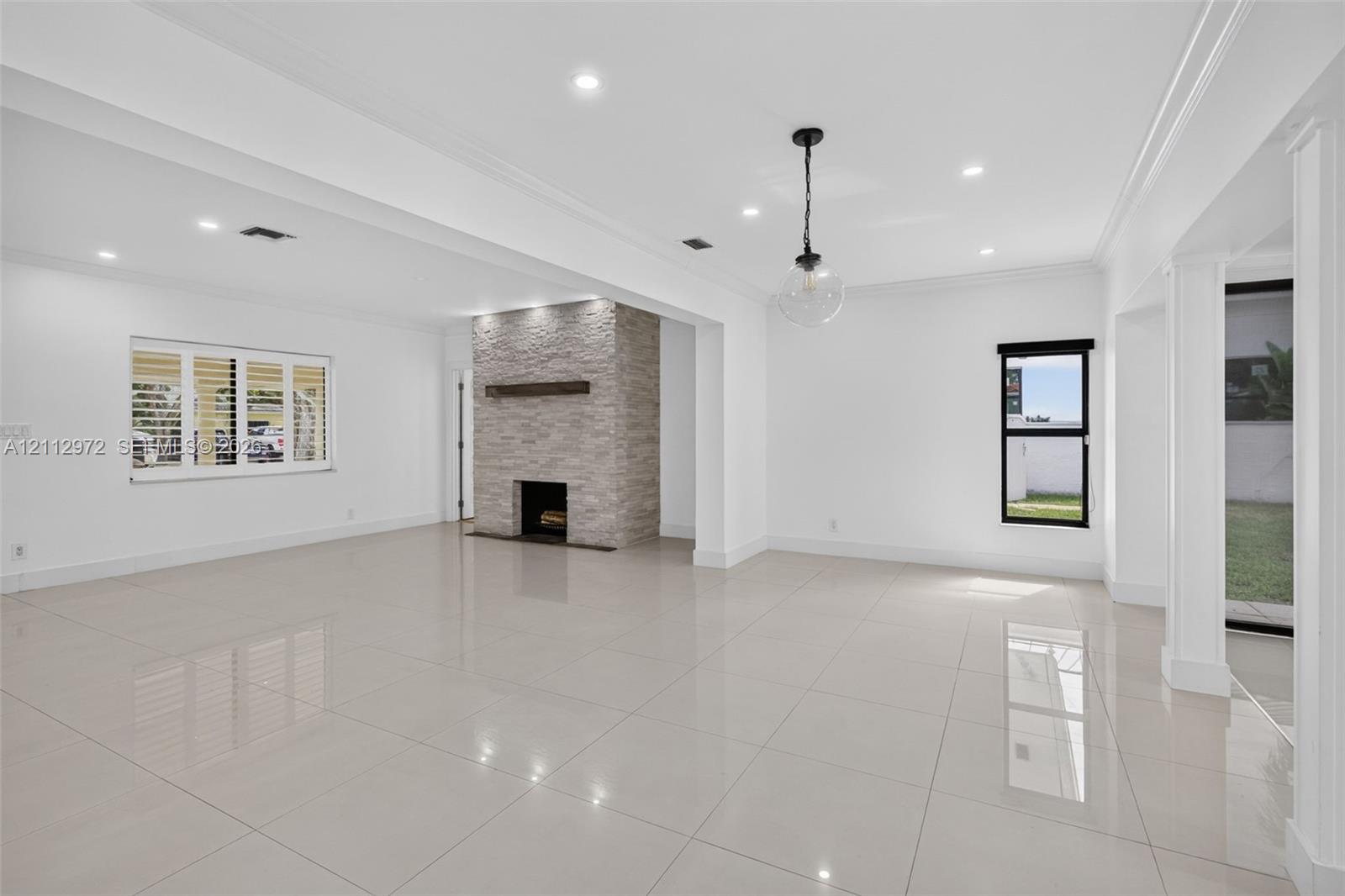 Open-concept interior featuring light-toned tile flooring, a floor-to-ceiling stacked stone fireplace with a wood mantel, recessed lighting, and a modern glass pendant fixture