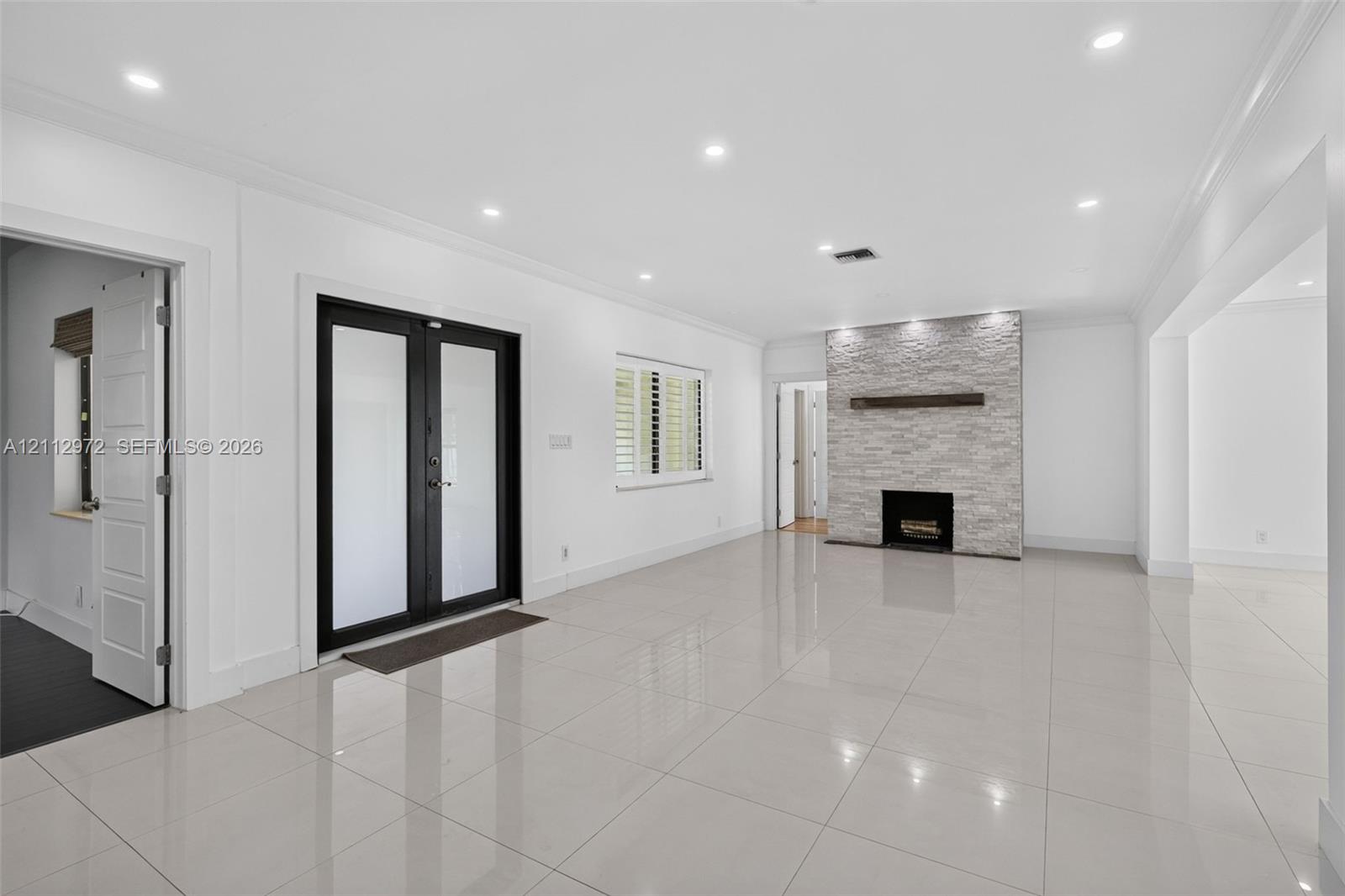 Large-format tile flooring, recessed ceiling lighting, stone-veneer fireplace with a wood mantel, and black-framed glass panel double doors Large-format tile flooring, recessed ceiling lighting, stone-veneer fireplace with a wood mantel, and black-framed glass panel double doors