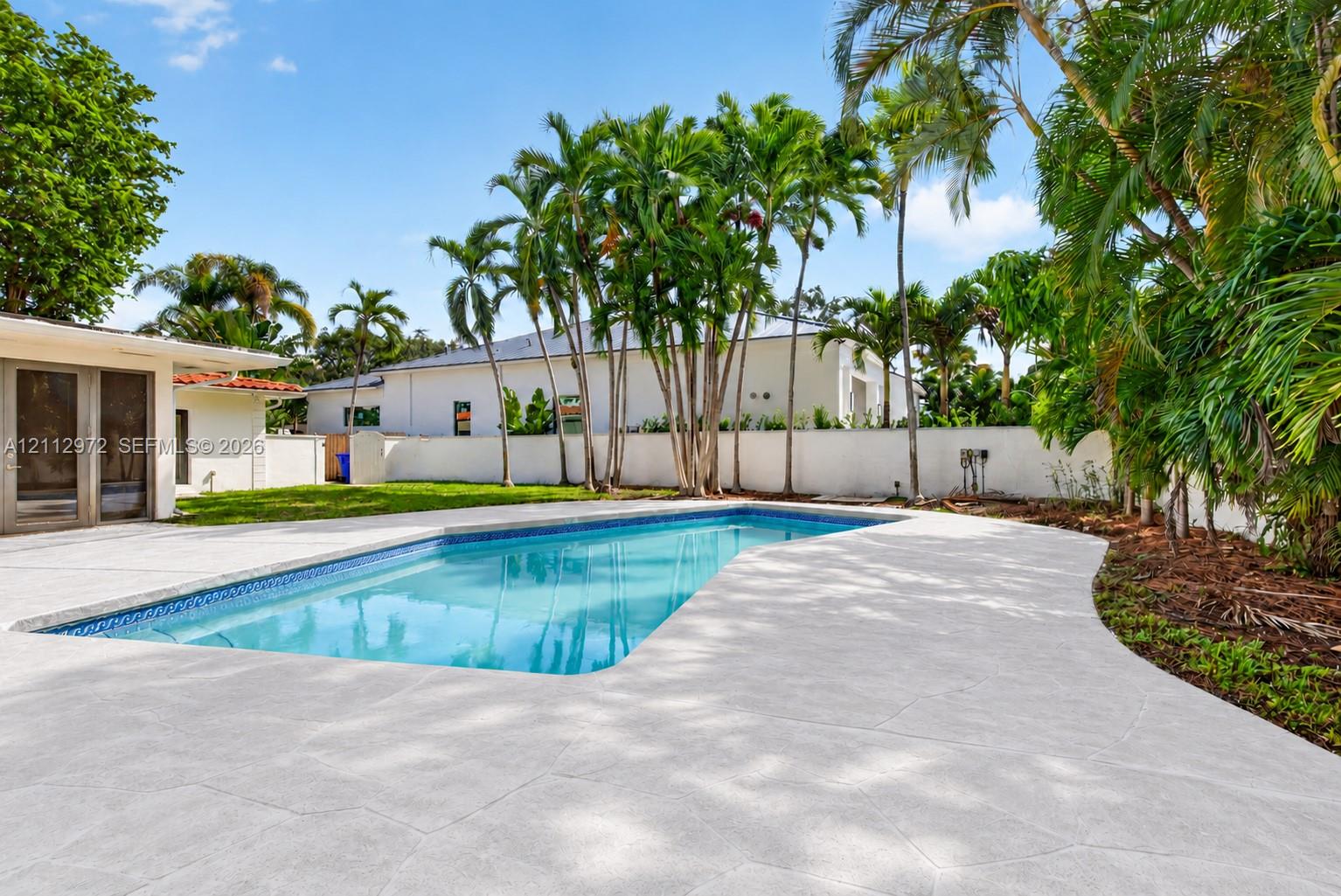 In-ground swimming pool featuring a textured concrete deck, privacy wall, manicured lawn, and mature palm trees