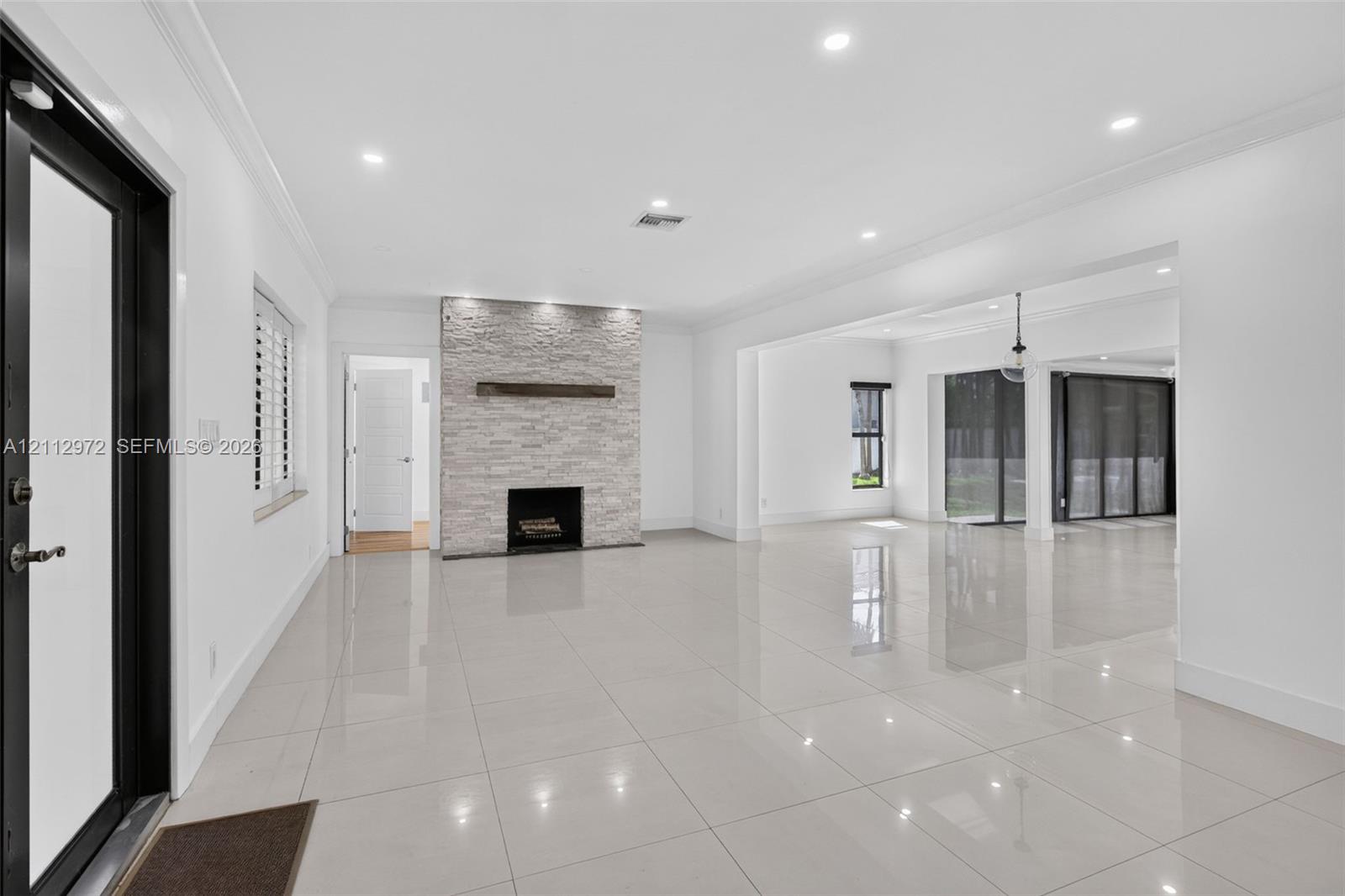Open-concept interior featuring large-format polished tile flooring, stone-veneer fireplace with a wood mantel, recessed ceiling lighting, and floor-to-ceiling glass sliding doors Open-concept interior featuring large-format polished tile flooring, stone-veneer fireplace with a wood mantel, recessed ceiling lighting, and floor-to-ceiling glass sliding doors