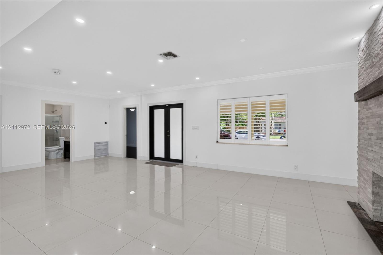 Large-format tile flooring, recessed ceiling lighting, stone-clad fireplace with a timber mantel, double-entry front door with glass insets, and windows featuring plantation shutters Large-format tile flooring, recessed ceiling lighting, stone-clad fireplace with a timber mantel, double-entry front door with glass insets, and windows featuring plantation shutters