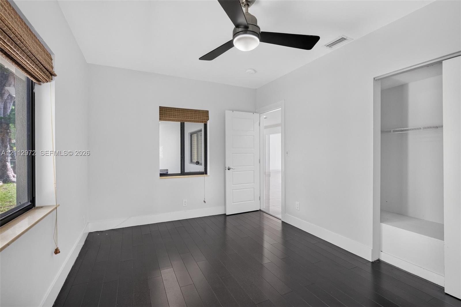 Room featuring dark wood-finish flooring, a contemporary matte black ceiling fan with integrated lighting, two windows with woven shades, and a built-in closet with sliding doors