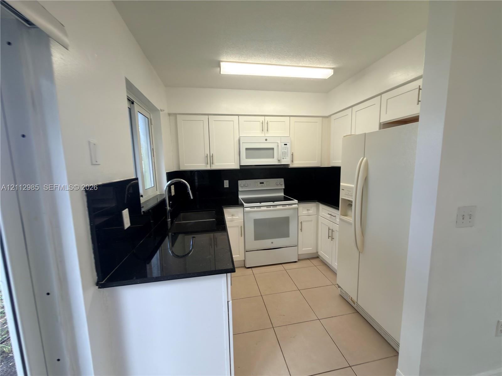 Kitchen featuring white cabinetry, black stone-finish countertops, black backsplash, white appliances, and tiled flooring Kitchen featuring white cabinetry, black stone-finish countertops, black backsplash, white appliances, and tiled flooring