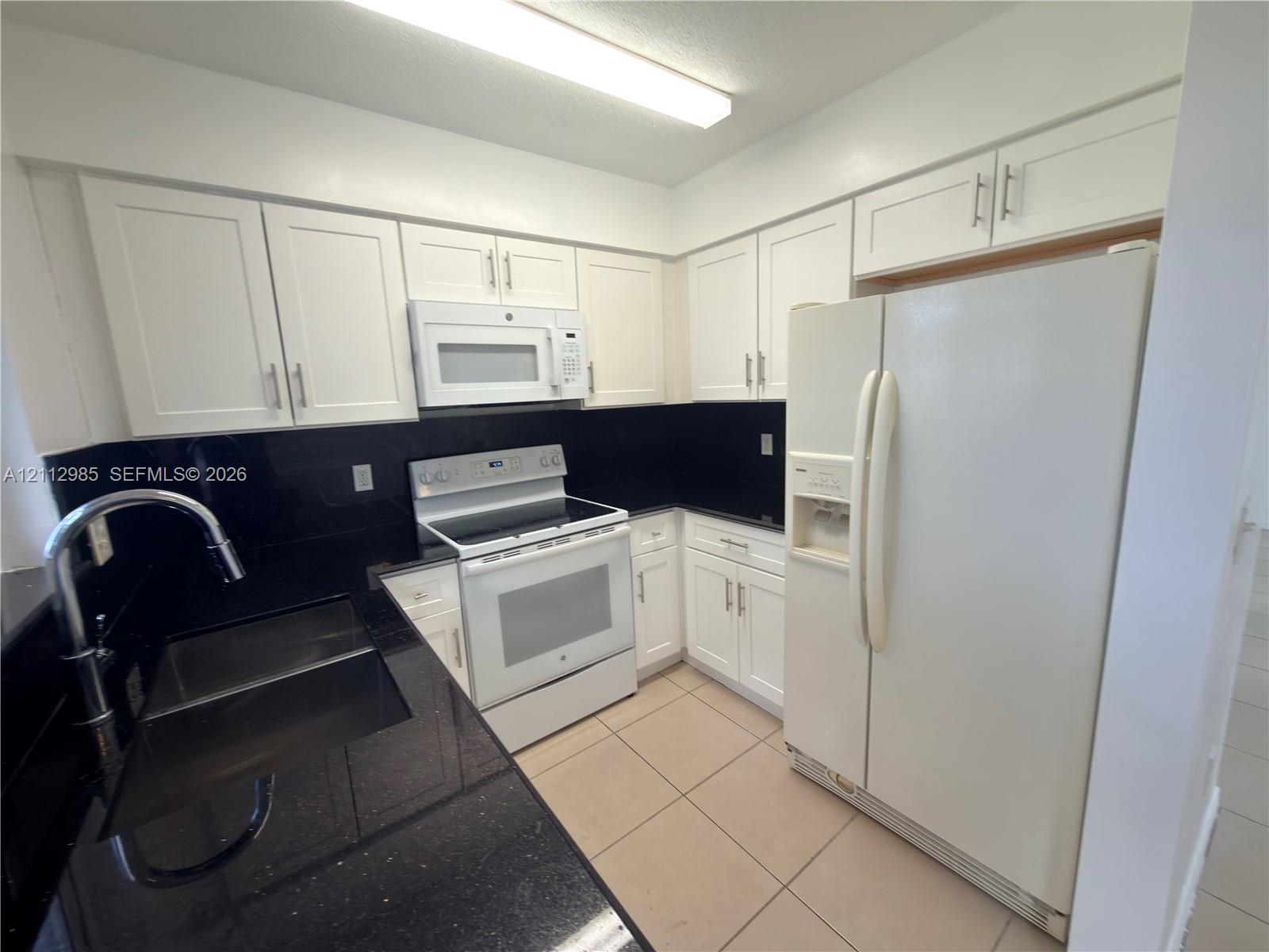 Kitchen featuring white cabinetry, black solid-surface countertops, integrated white appliances, and tiled flooring Kitchen featuring white cabinetry, black solid-surface countertops, integrated white appliances, and tiled flooring