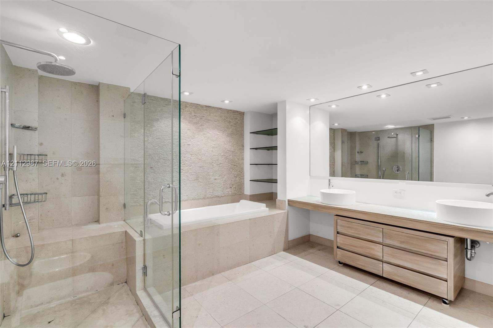 Floating wood-finish vanity with dual vessel sinks, glass-enclosed shower, built-in soaking tub, textured stone accent wall, and expansive frameless vanity mirror