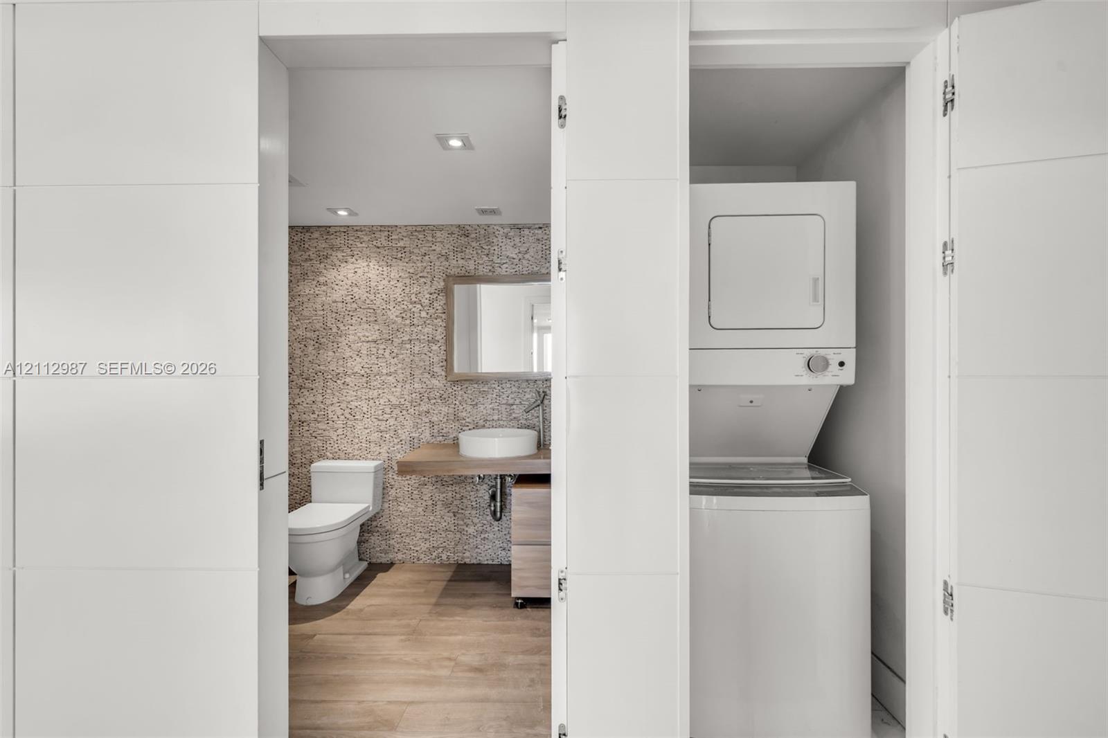 Stacked laundry unit set within a custom closet, featuring a floating vanity with a vessel sink, textured stone-tiled wall, and wood-finish flooring