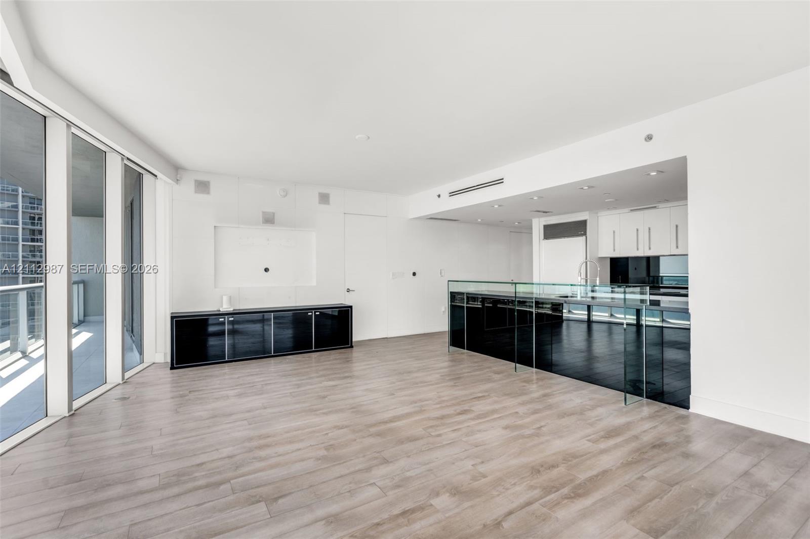 Open-concept floor plan featuring wood-finish flooring, floor-to-ceiling glass wall, built-in black lacquer cabinetry, and a kitchen island with a glass countertop Open-concept floor plan featuring wood-finish flooring, floor-to-ceiling glass wall, built-in black lacquer cabinetry, and a kitchen island with a glass countertop
