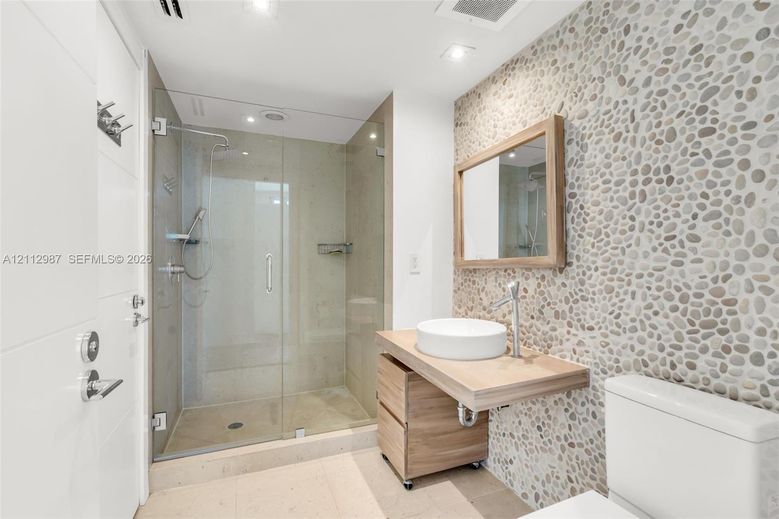 Bathroom featuring a pebble-tiled accent wall, floating wood-finish vanity with a white vessel sink, and a walk-in glass-enclosed shower with polished chrome fixtures
