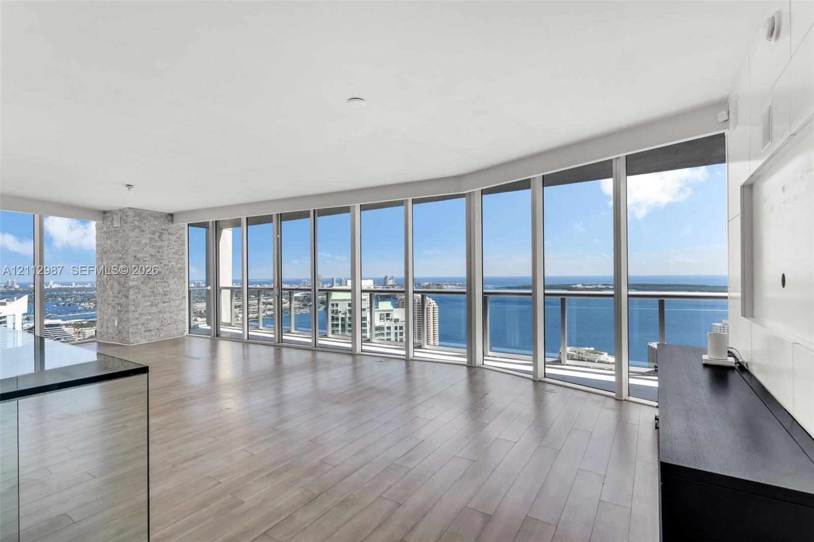 Floor-to-ceiling windows, wood-finish flooring, stone-veneer accent pillar, glass-panel balcony railing, and expansive water backdrop Floor-to-ceiling windows, wood-finish flooring, stone-veneer accent pillar, glass-panel balcony railing, and expansive water backdrop