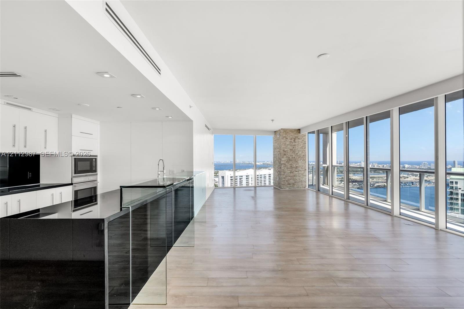 Open-concept interior featuring floor-to-ceiling windows, wood-finish flooring, a kitchen with black countertops and white cabinetry, and a stone-clad accent pillar Open-concept interior featuring floor-to-ceiling windows, wood-finish flooring, a kitchen with black countertops and white cabinetry, and a stone-clad accent pillar
