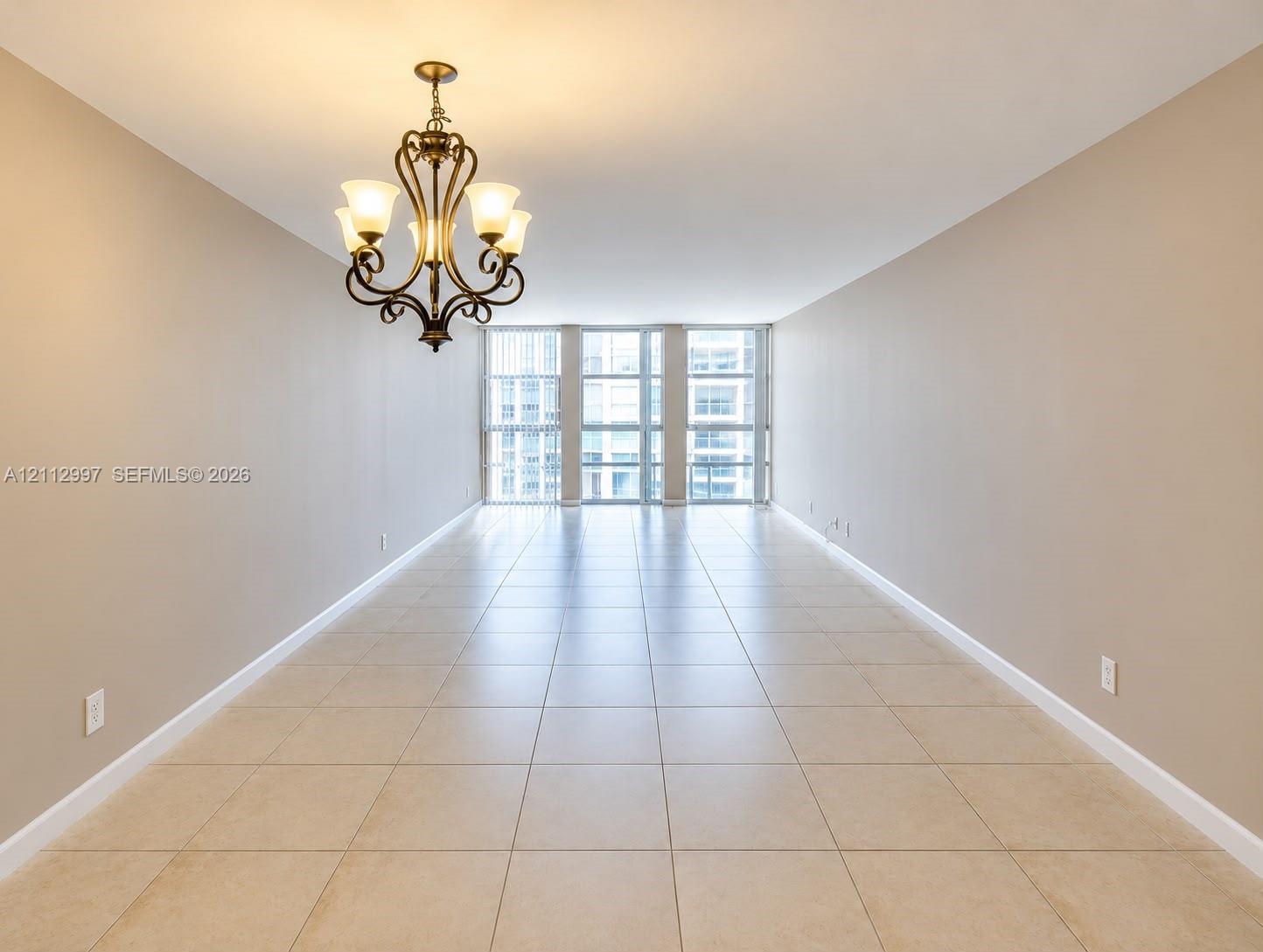 Spacious room featuring neutral-toned tile flooring, floor-to-ceiling windows, and a decorative multi-light metal chandelier Spacious room featuring neutral-toned tile flooring, floor-to-ceiling windows, and a decorative multi-light metal chandelier