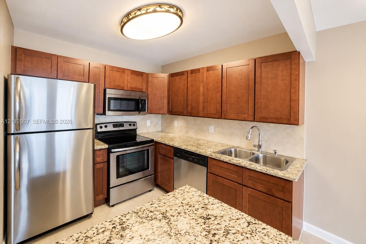 Kitchen featuring wood cabinetry, stone countertops, stainless steel appliances, a double-basin undermount sink with a gooseneck faucet, and a decorative flush-mount ceiling fixture Kitchen featuring wood cabinetry, stone countertops, stainless steel appliances, a double-basin undermount sink with a gooseneck faucet, and a decorative flush-mount ceiling fixture