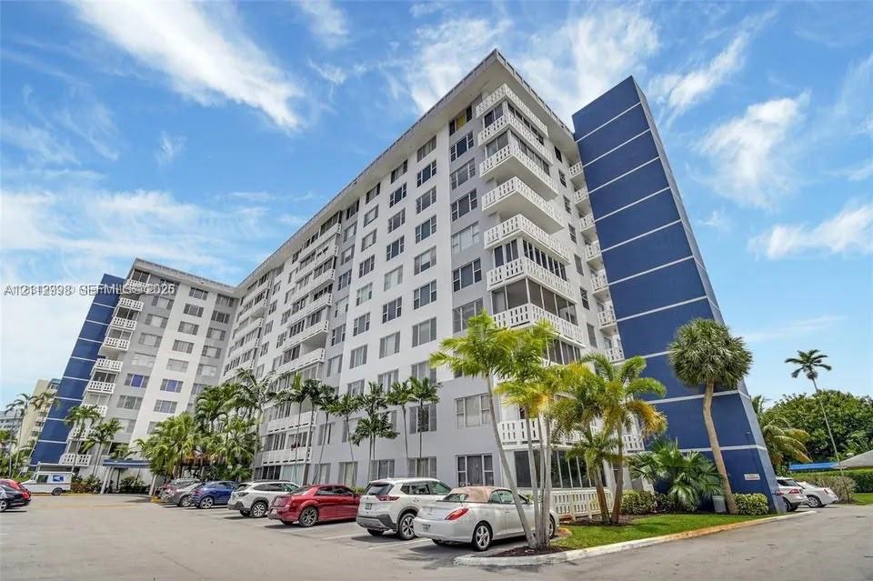 Multi-story residential building featuring a blue and white facade, private railed balconies, and surface parking