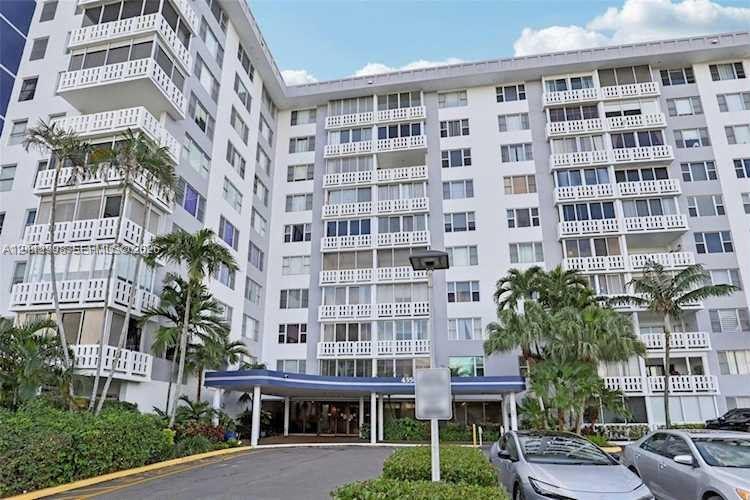 High-rise building exterior featuring structural balcony enclosures, decorative latticed railings, covered entryway portico, and mature palm landscaping