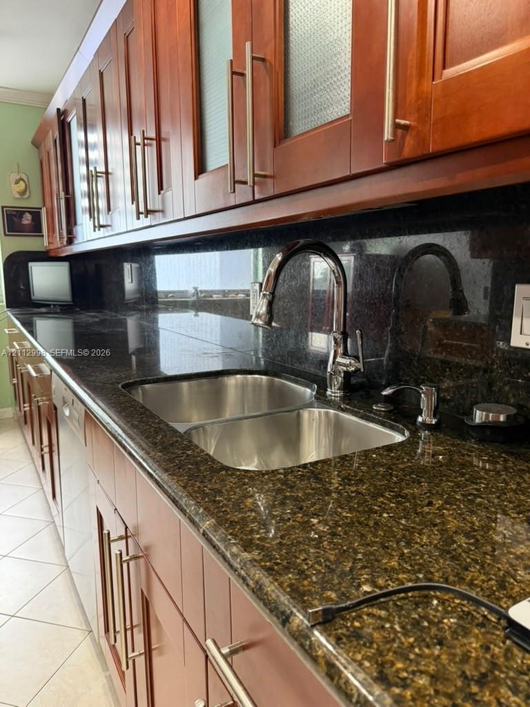 Kitchen featuring warm-toned wood cabinetry, dark stone countertops, an integrated stainless steel double-basin sink, a gooseneck faucet, and tiled flooring
