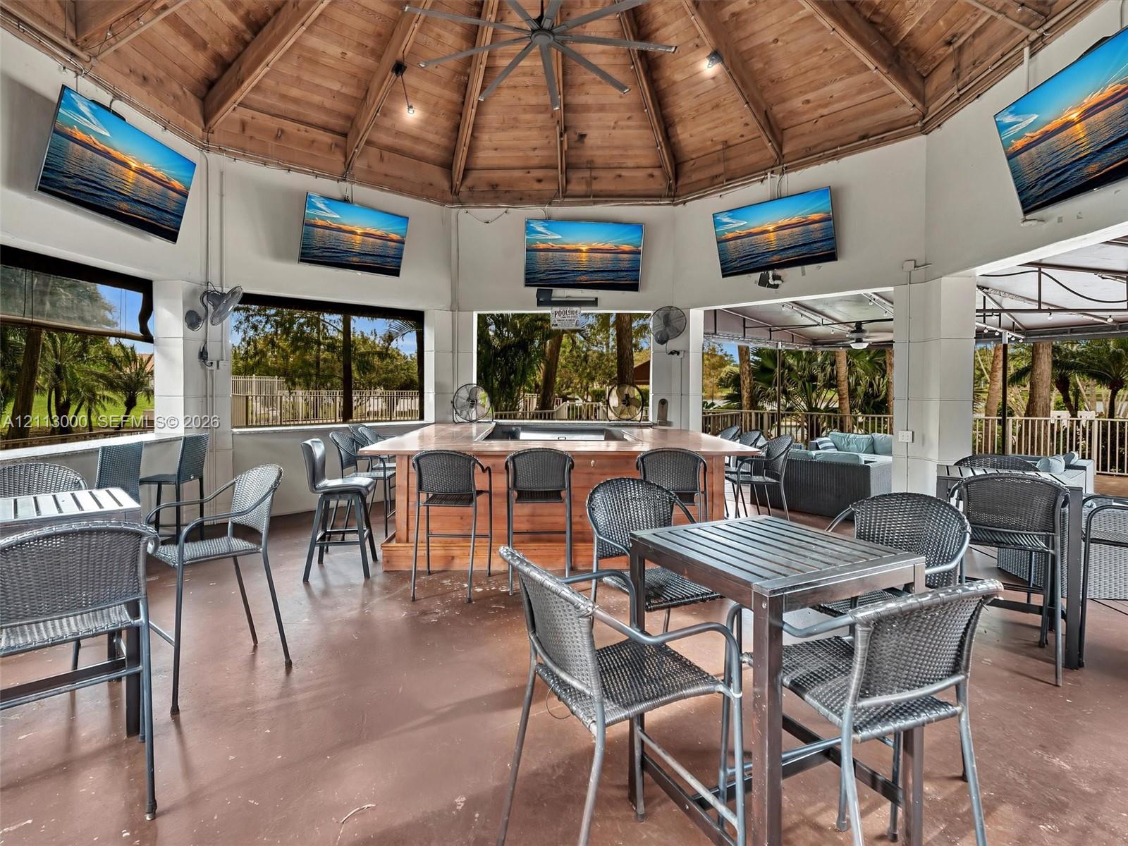 Covered outdoor pavilion featuring a vaulted wood-beam ceiling, built-in center bar, stone-finish flooring, and multiple wall-mounted television screens