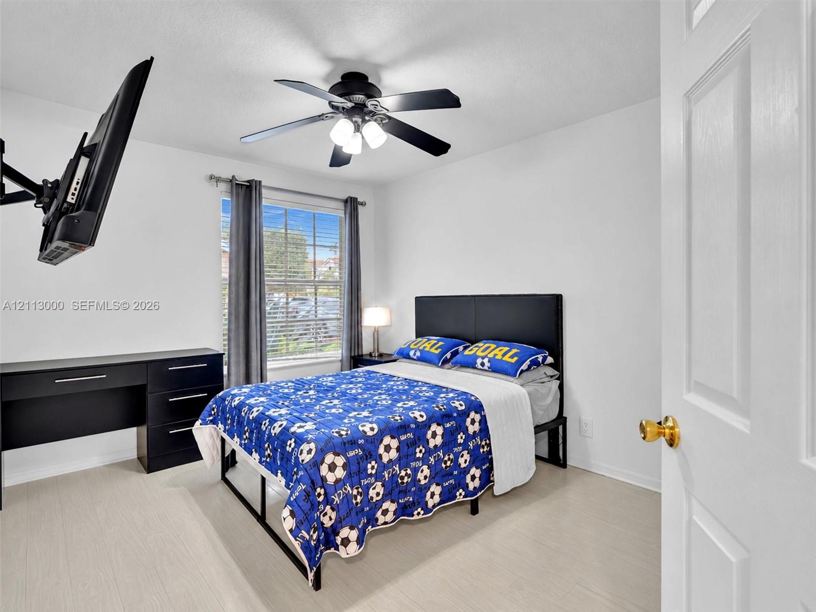 Room featuring wood-finish flooring, a matte black multi-blade ceiling fan with integrated lighting, a dual-pane window, and a multi-panel door with brass-finish hardware
