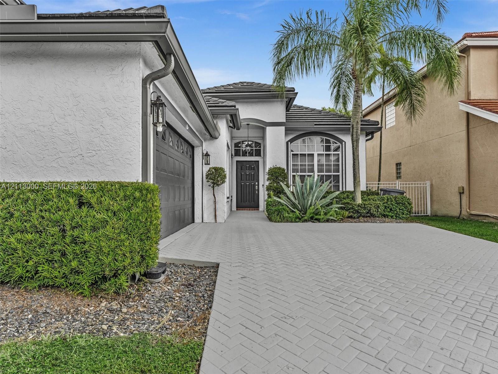 Stucco exterior featuring a herringbone-patterned paver driveway, arched entryway, dark-tone garage door, and decorative transom window