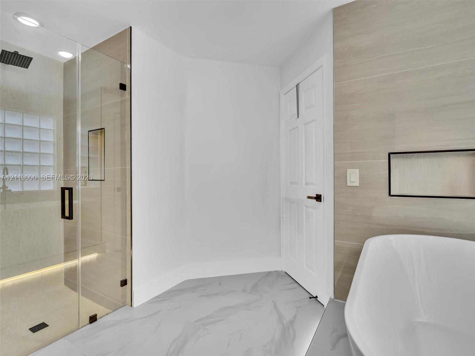 Walk-in shower with frameless glass enclosure, rainfall showerhead, and integrated recessed shelving, featuring stone-finish flooring and a freestanding soaking tub