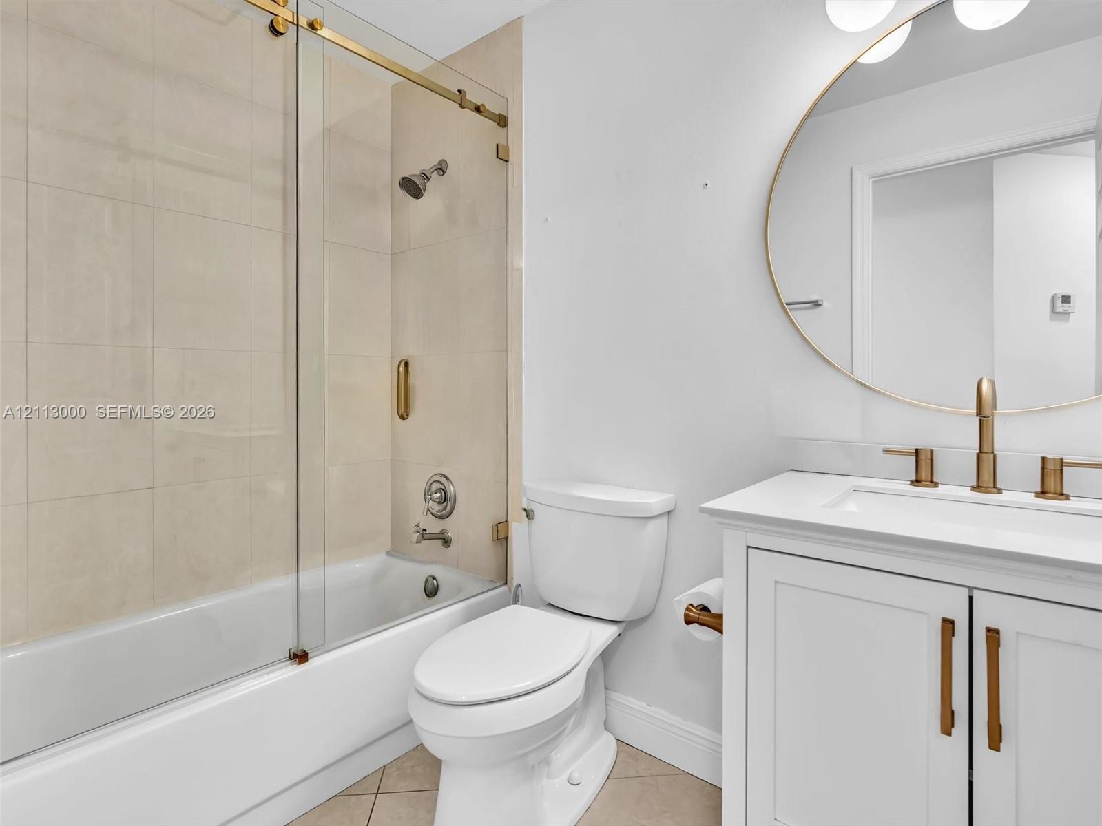 Bathroom featuring a white vanity cabinet with a stone countertop, an oval mirror with gold-tone trim, gold-tone faucet and hardware, and a glass-enclosed tub shower with tile surround