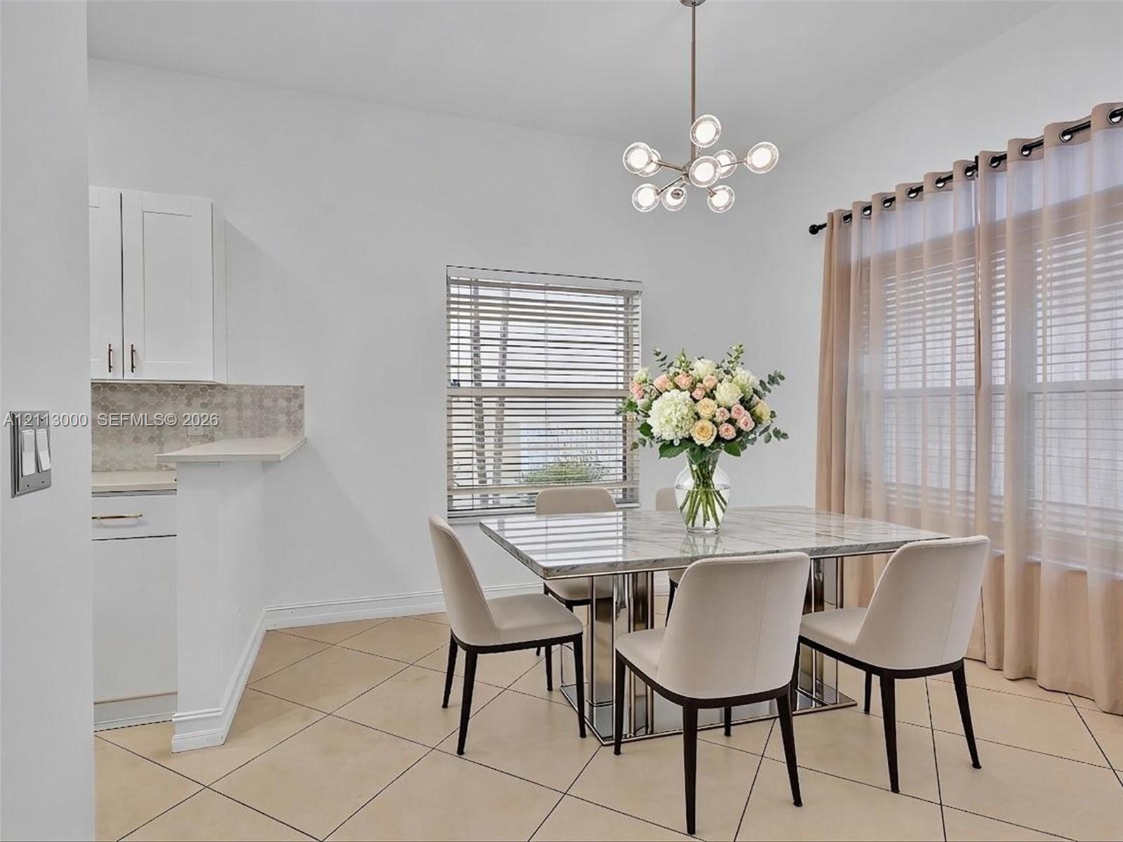 Neutral-toned tile flooring, modern multi-globe chandelier, white shaker-style cabinetry, and large windows with horizontal blinds