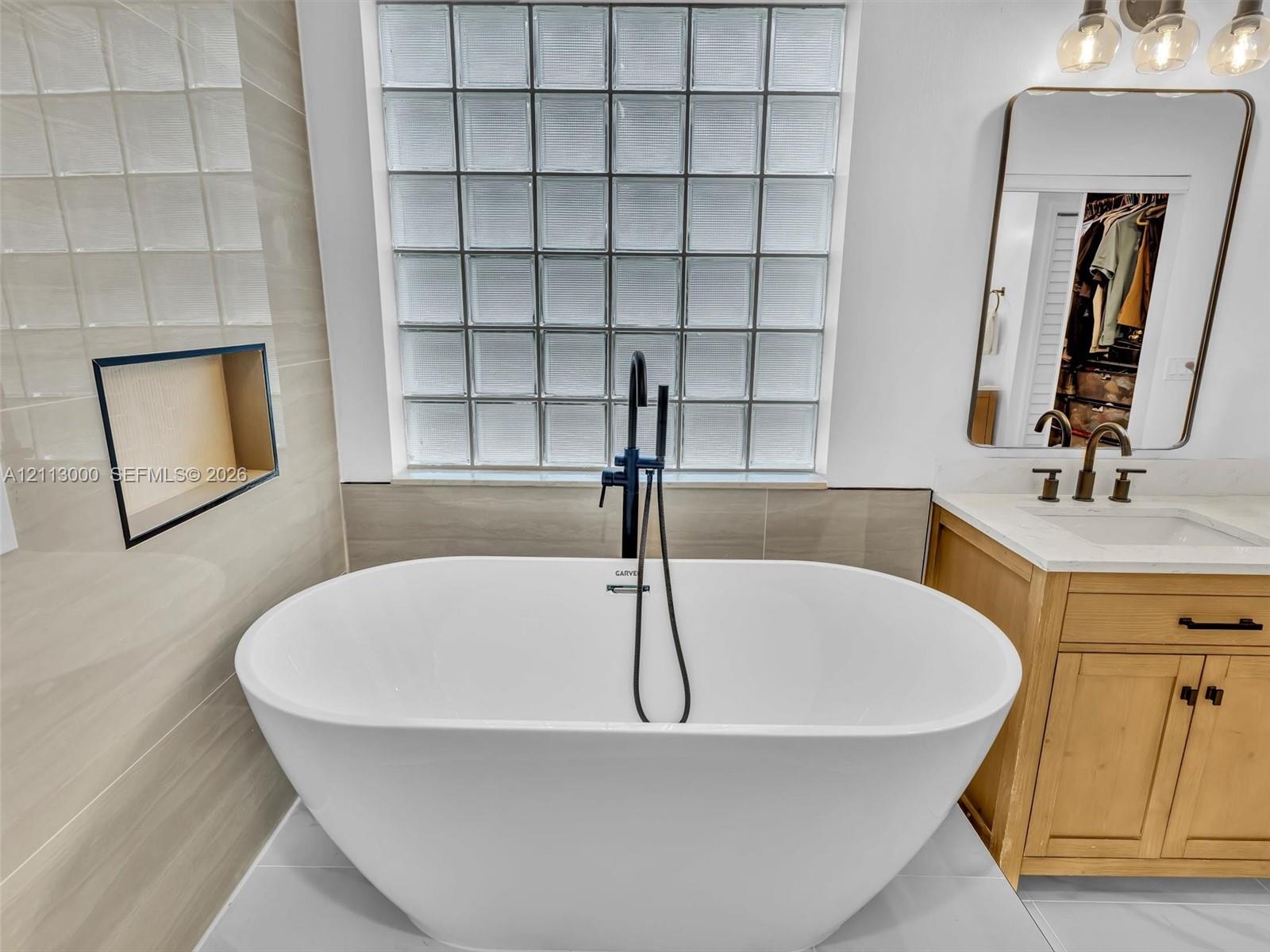 Freestanding soaking tub featuring a matte black floor-mounted faucet with hand shower, glass block window, light-toned wood vanity with integrated countertop, and recessed tiled wall niche