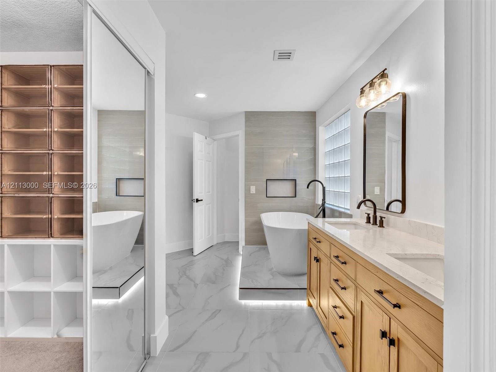 Bathroom featuring a dual-sink vanity with wood-finish cabinetry, a freestanding soaking tub set on a raised platform, built-in floor-to-ceiling shelving, large-format tile flooring, and glass block window detailing