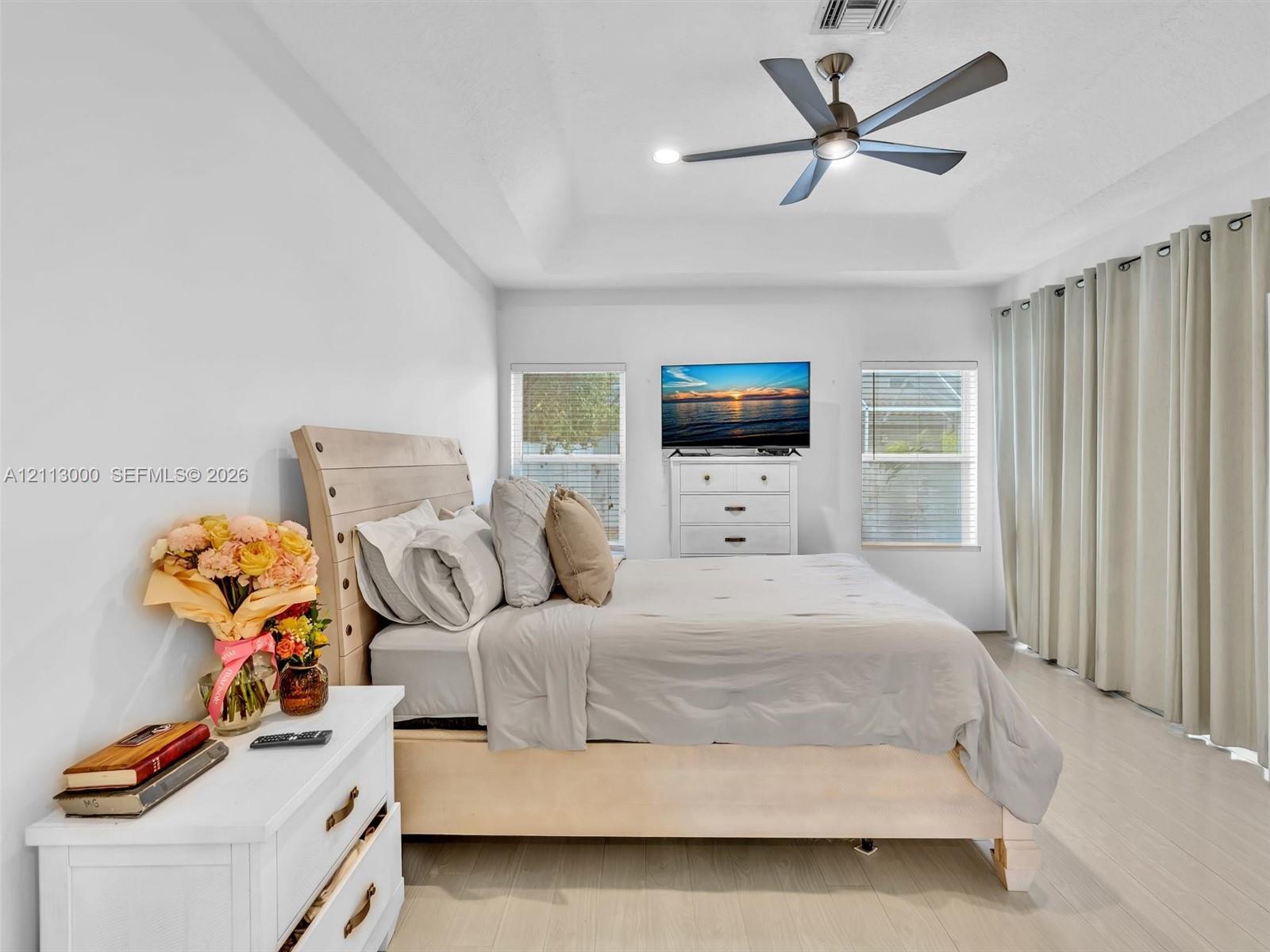 Tray ceiling, multi-blade ceiling fan with integrated lighting, wood-finish flooring, and large windows with full-length window treatments