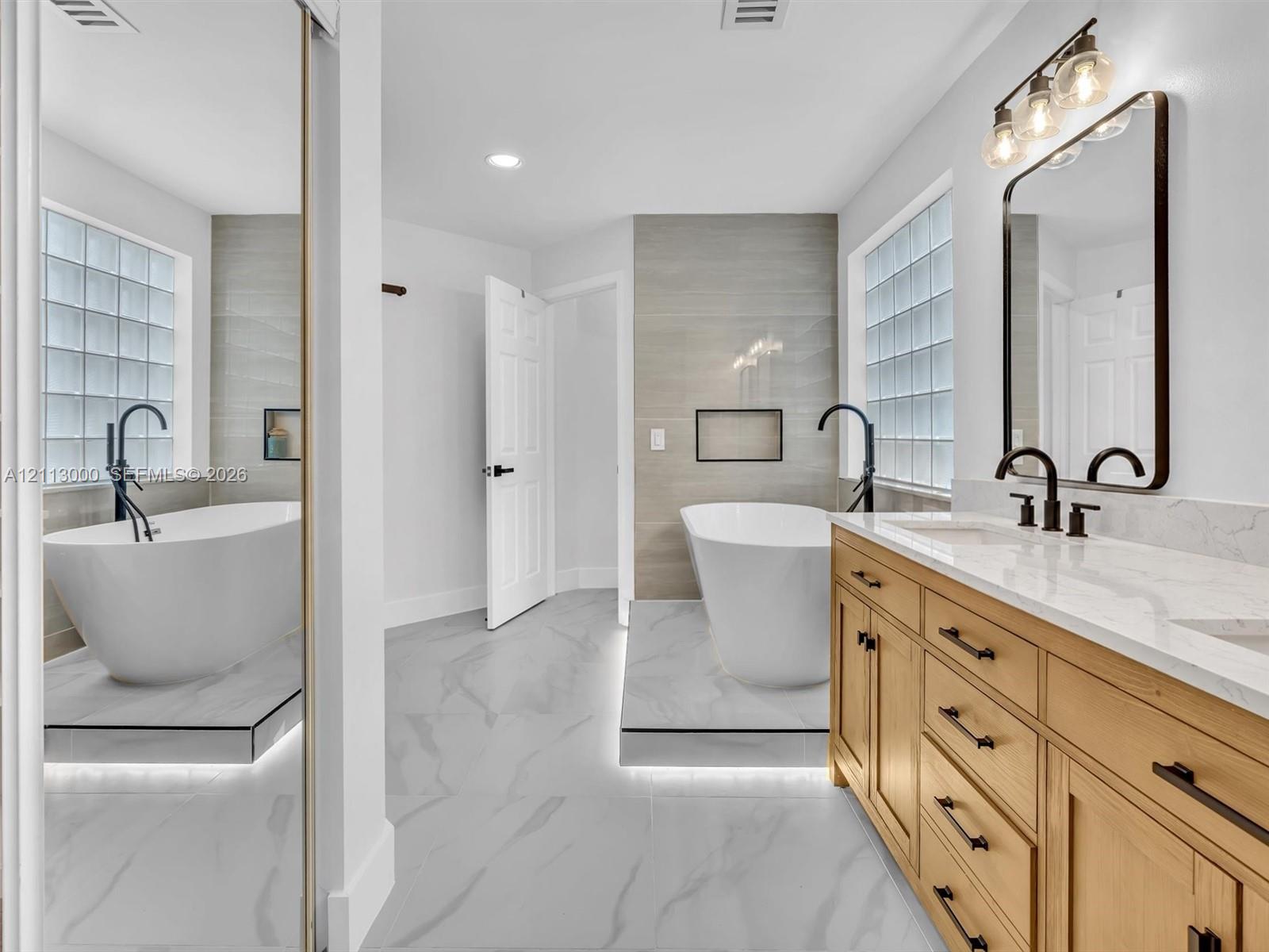 Bathroom featuring a freestanding soaking tub on a raised platform with under-cabinet lighting, wood-finish cabinetry, quartz countertop with dual integrated sinks, and glass block windows