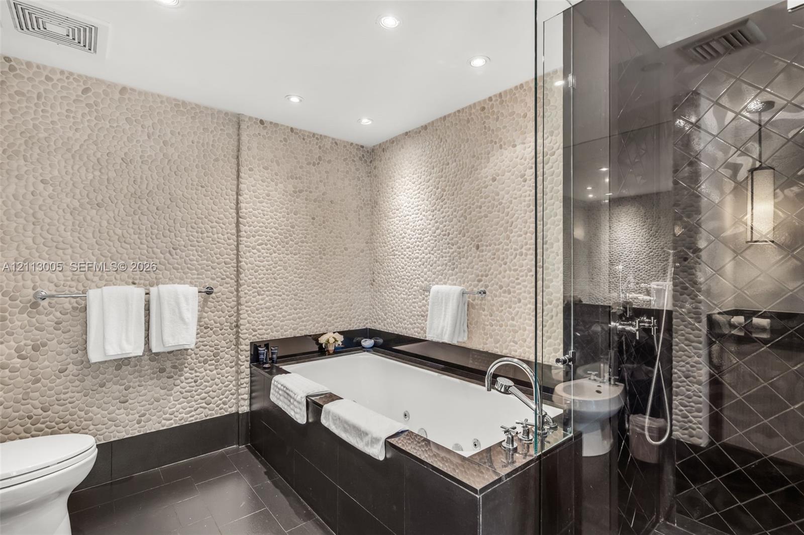 Bathroom featuring pebble-textured stone accent walls, a jetted soaking tub with a dark stone surround, a glass-enclosed shower with diamond-patterned tile, and recessed ceiling lighting