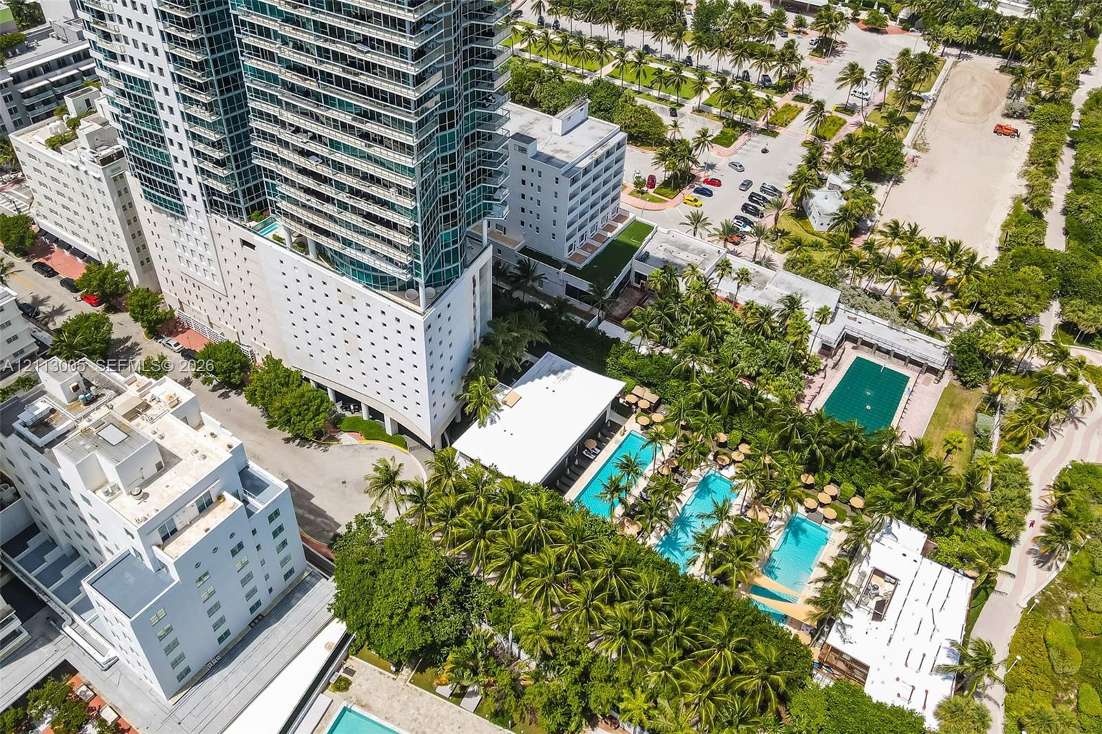 High-rise residential structure featuring glass-paneled balconies, multiple swimming pools, extensive lush landscaping with mature palm trees, and hardscaped courtyard areas
