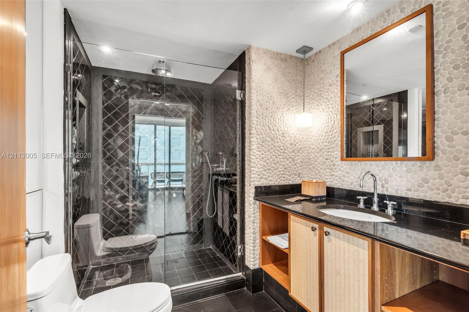 Bathroom featuring a pebble-stone accent wall, custom wood-finish vanity with a dark countertop, glass-enclosed shower with black diamond-patterned tile, and chrome fixtures