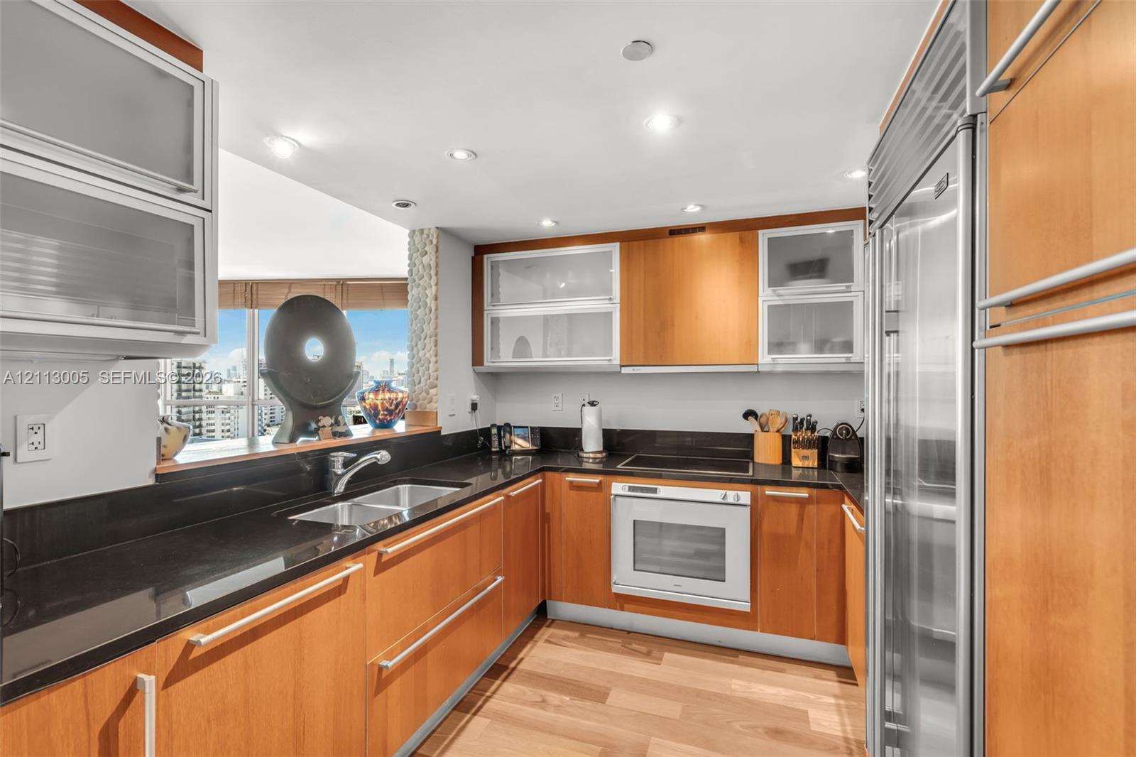 Kitchen featuring wood-veneer cabinetry with frosted glass inserts, dark solid-surface countertops, built-in stainless steel refrigerator, recessed lighting, and wood-finish flooring