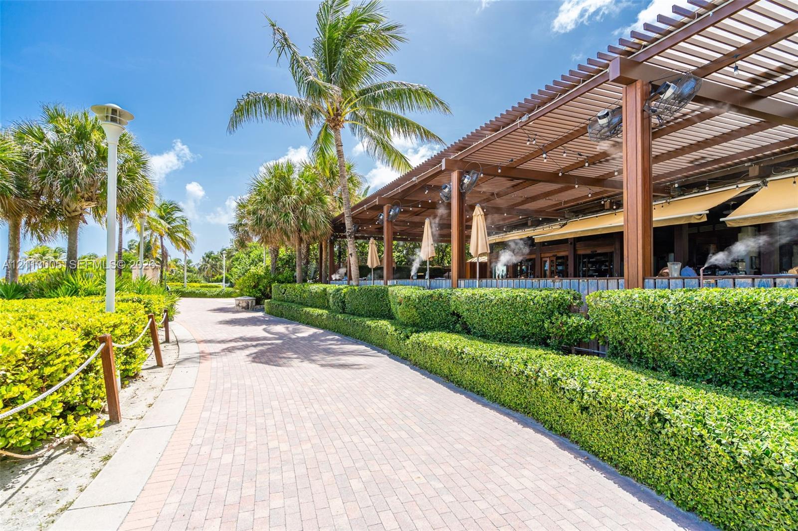 Outdoor paver walkway bordered by manicured hedging and palm trees, featuring an expansive wood-frame pergola with mounted outdoor cooling fans