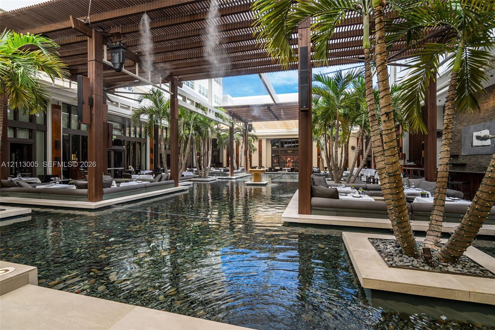Expansive courtyard featuring a reflection pool, overhead wood-slat pergola, integrated palm tree planters, stone tile hardscaping, and built-in masonry seating